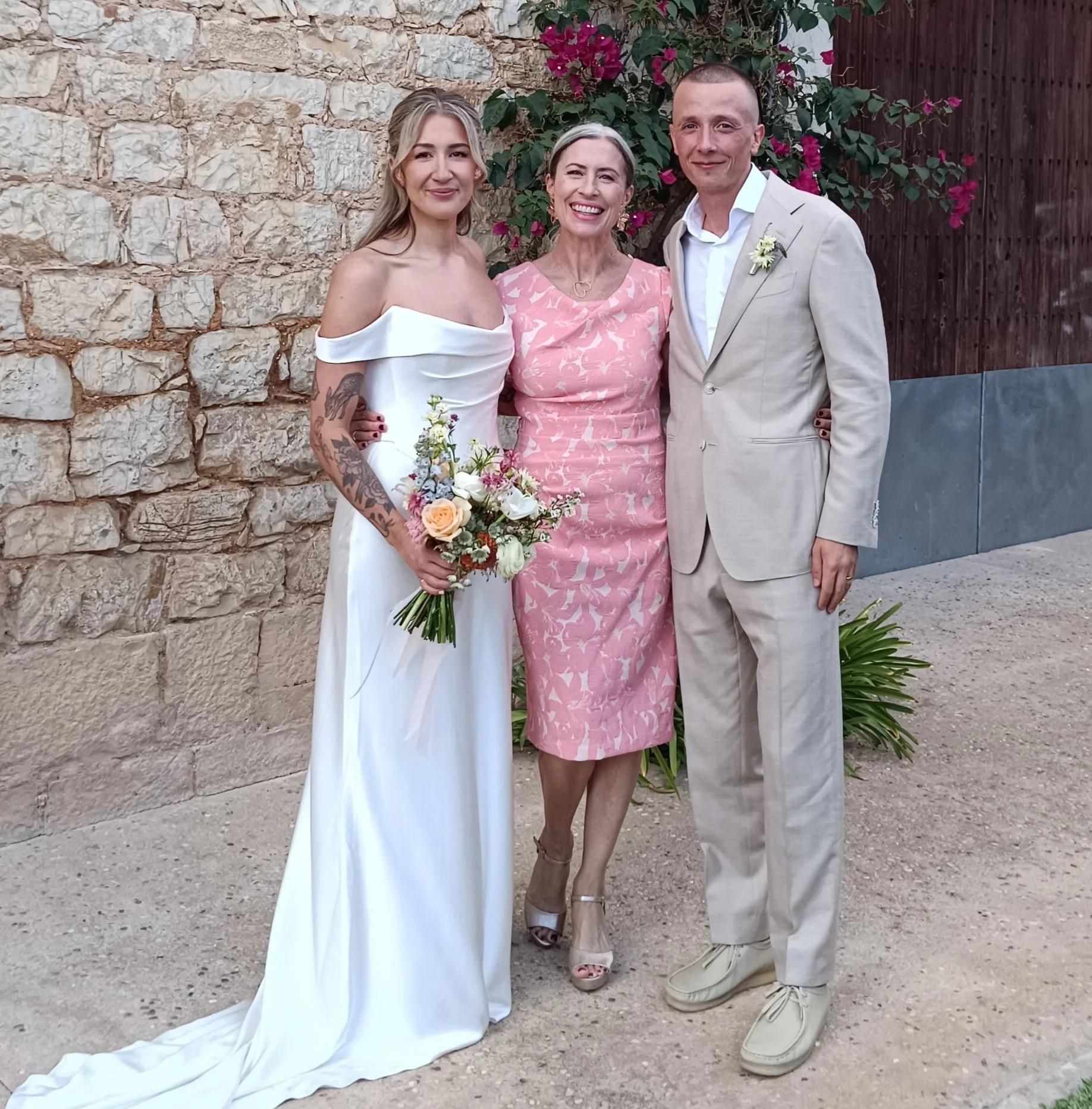 Three people standing outdoors against a stone wall with pink flowers, dressed in wedding attire, smiling at the camera.
