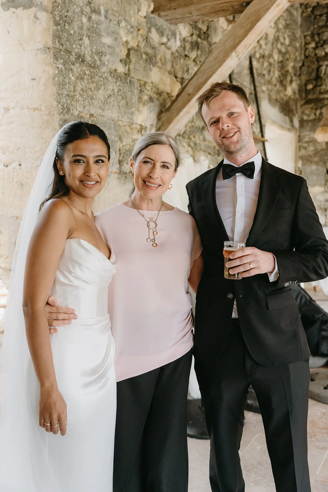 Three people at a wedding reception: a bride in a white wedding dress, an older woman in a light pink blouse, and a groom in a black tuxedo holding a drink, smiling in a rustic setting with stone walls.