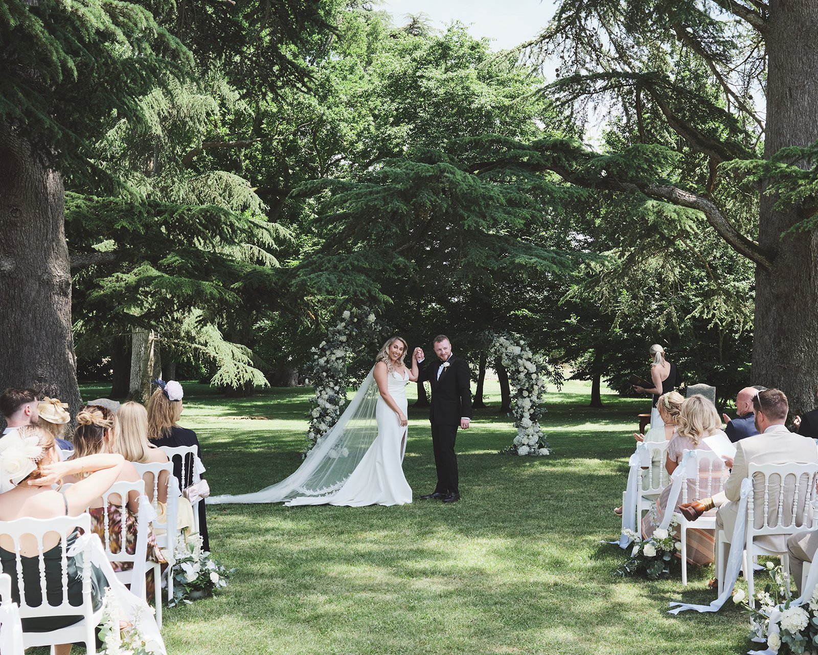 An outdoor wedding ceremony with a bride and groom holding hands under a floral arch, surrounded by seated guests, in a lush green park with large trees.
