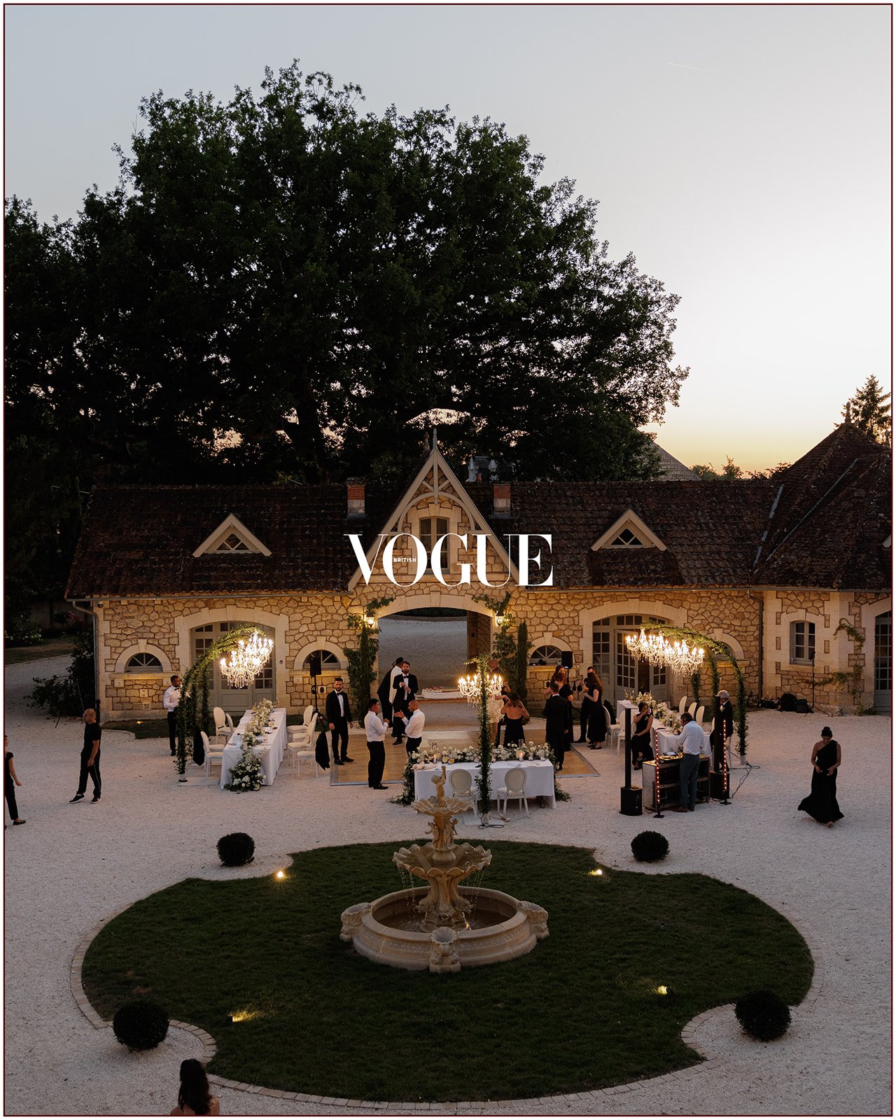 An evening outdoor wedding reception with a stone building, string lights, and guests mingling in formal attire, surrounding a fountain and lush landscaped area.