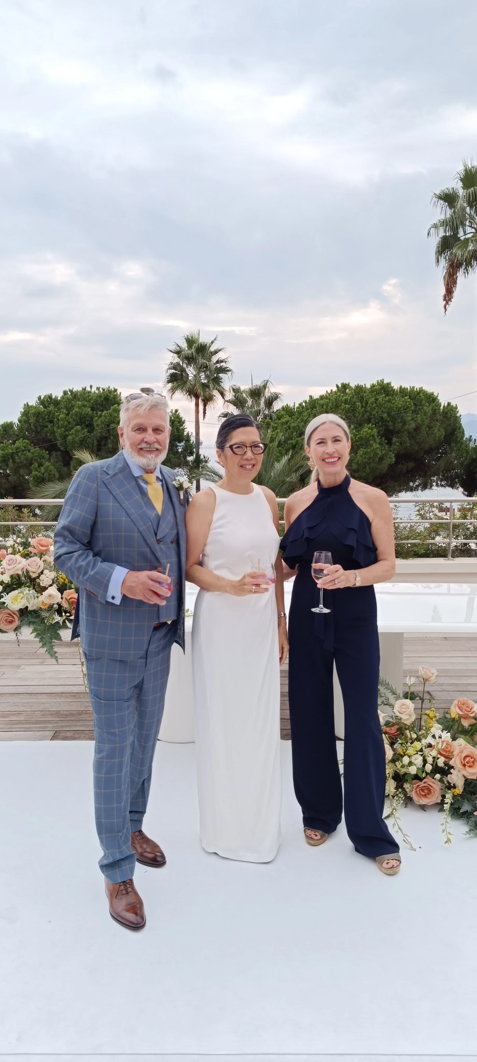 Three people at a wedding in Cannes performed by Laura the Chateau Celebrant standing on a white platform with floral arrangements, holding glasses of wine, outdoors with trees and cloudy sky in the background.
