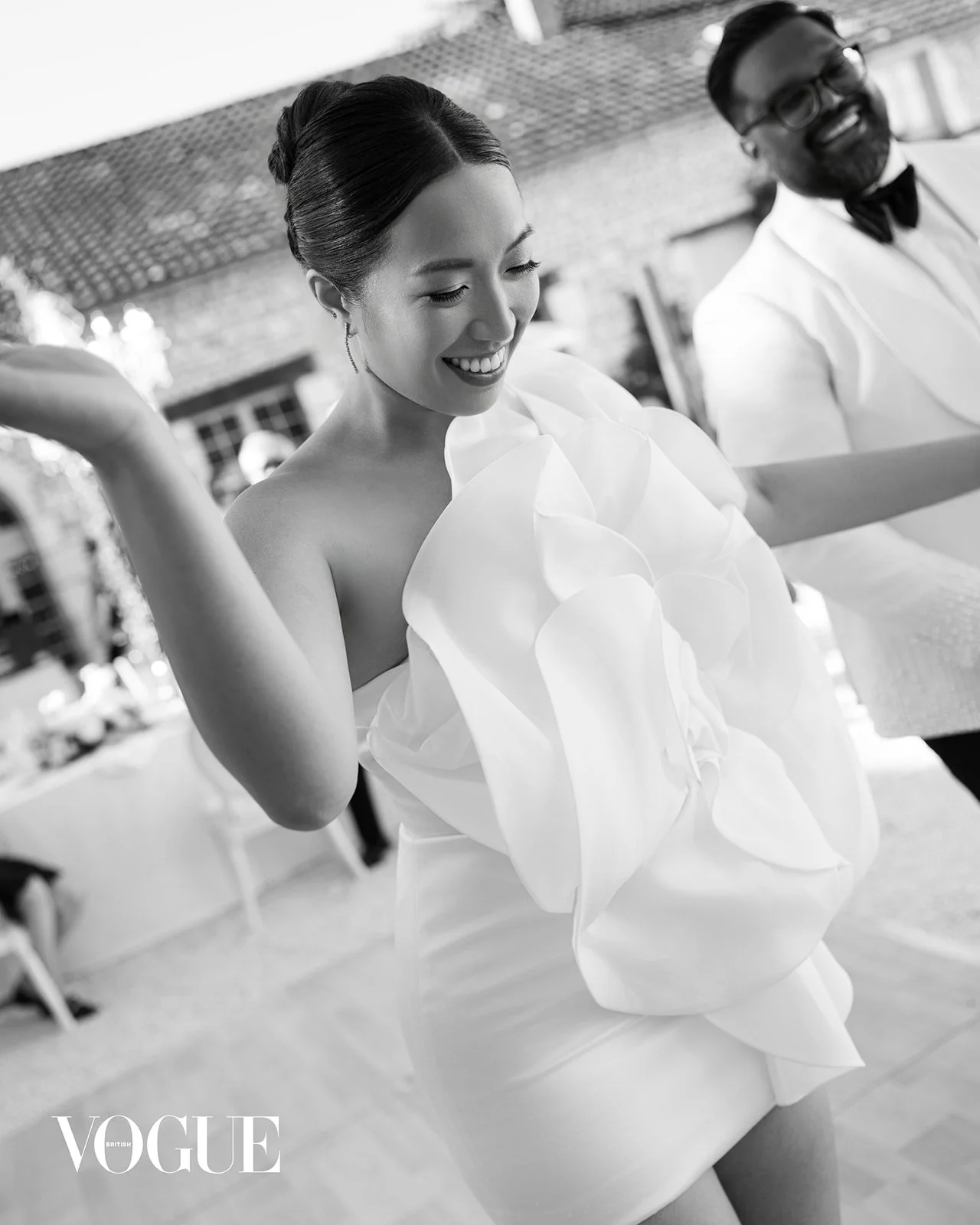 A woman in an elegant dress, smiling, with a man in a tuxedo in the background, at an outdoor event. The image is in black and white, with a "VOGUE" logo in the bottom left corner.