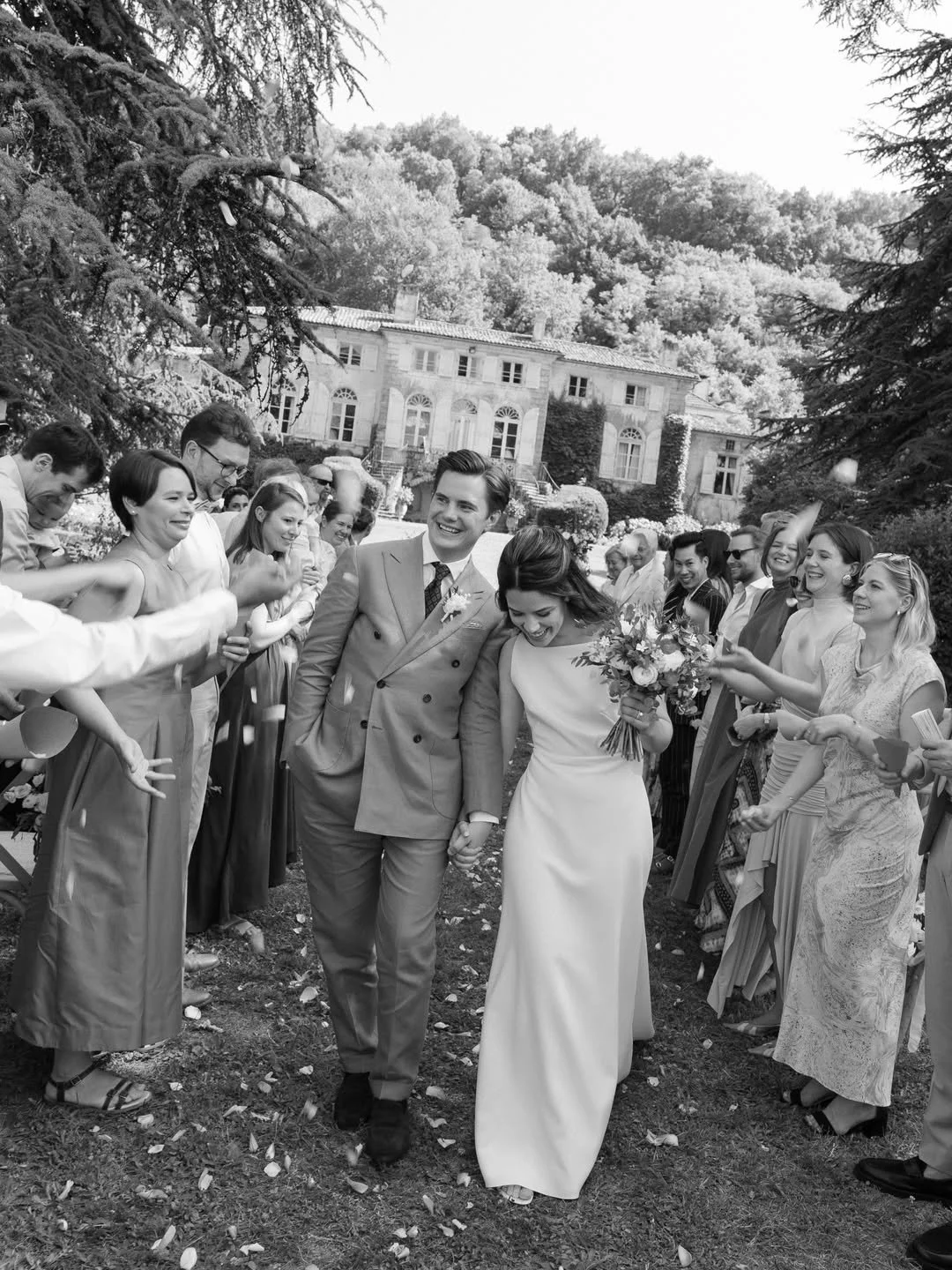 A black-and-white photo of a bride and groom walking hand-in-hand through a crowd of friends and family at an outdoor wedding, surrounded by trees and a large historic house in the background.