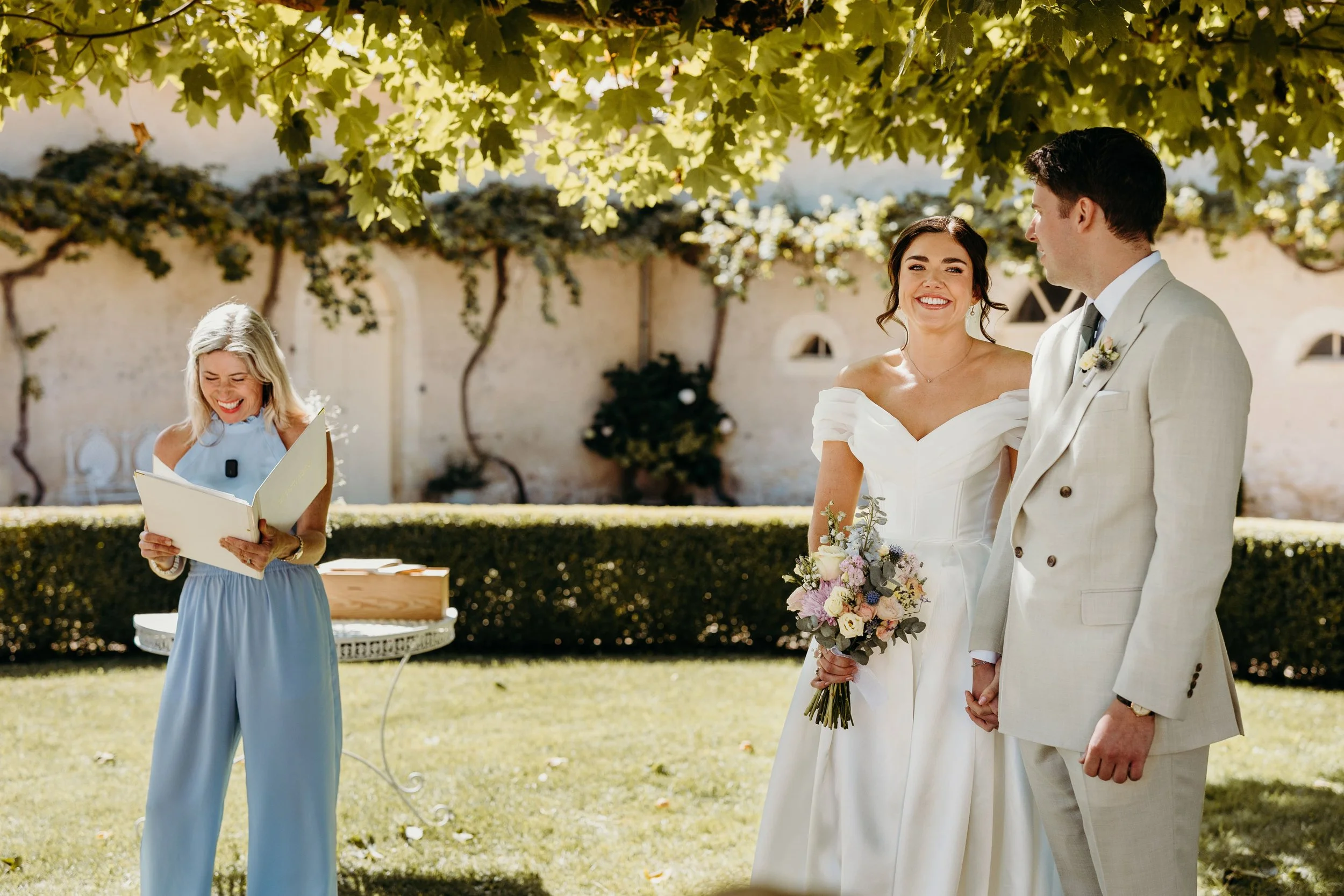 A bride and groom holding hands and smiling during their outdoor wedding ceremony, with an officiant reading vows beside them under a canopy of green leaves. At Chateau Gassies.