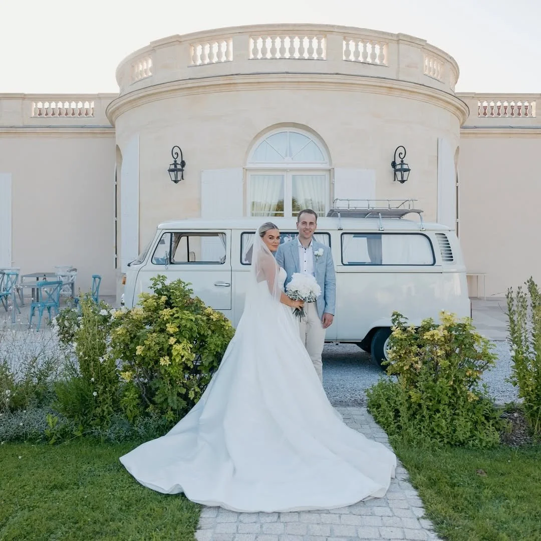 Bride and groom standing in front of a vintage white van on a wedding day, with a grand building in the background and lush greenery surrounding them.