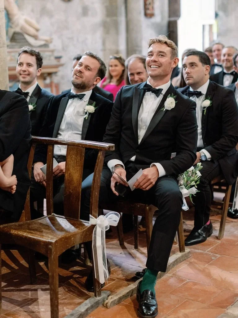 A group of men dressed in black tuxedos with white shirts and black bow ties sitting in pews during a wedding ceremony. One man in the front row is smiling and holding a paper, with a bouquet of flowers on his chair, inside a church with stone walls and religious statues.