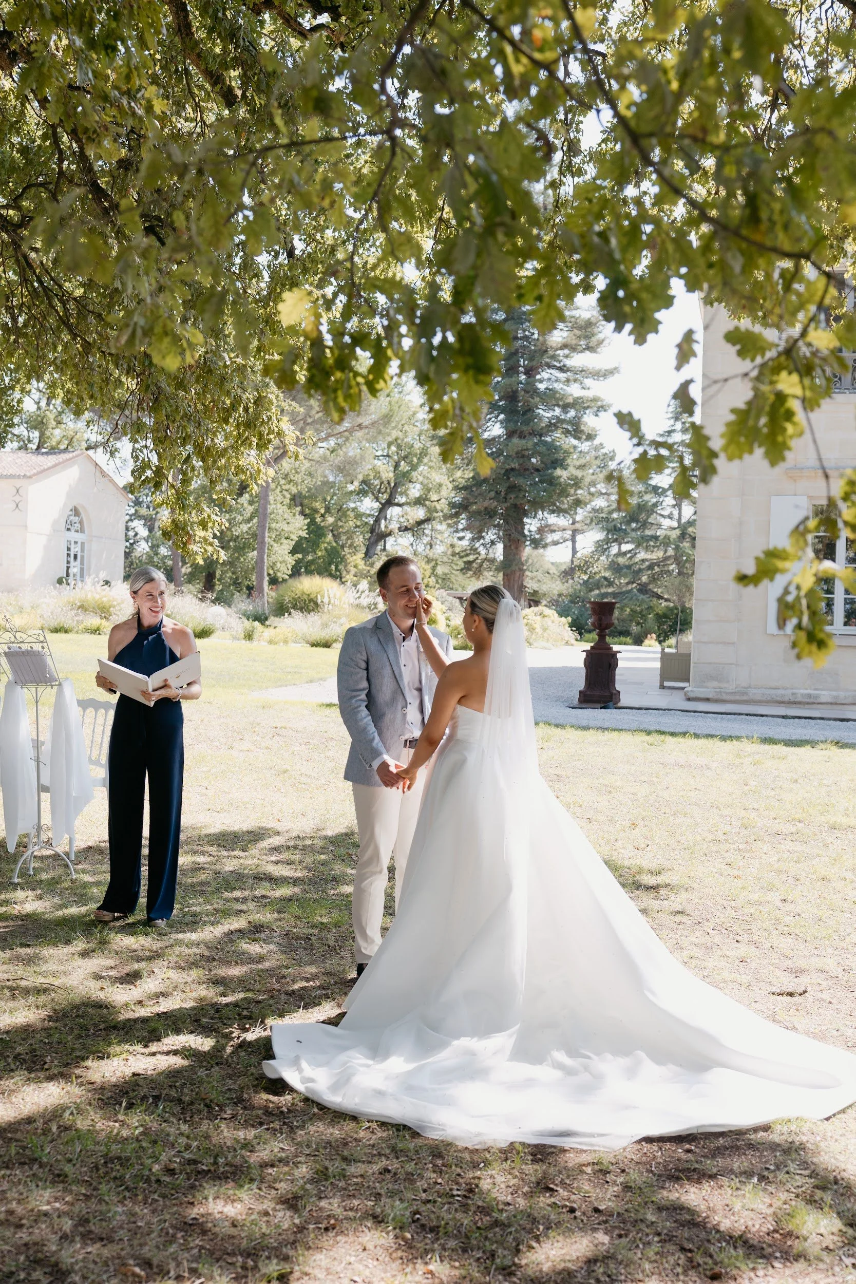 A bride and groom holding hands, standing outdoors under a tree, exchanging vows during a wedding ceremony, with an officiant Laura , The Chateau Celebrant holding a book and smiling nearby.
