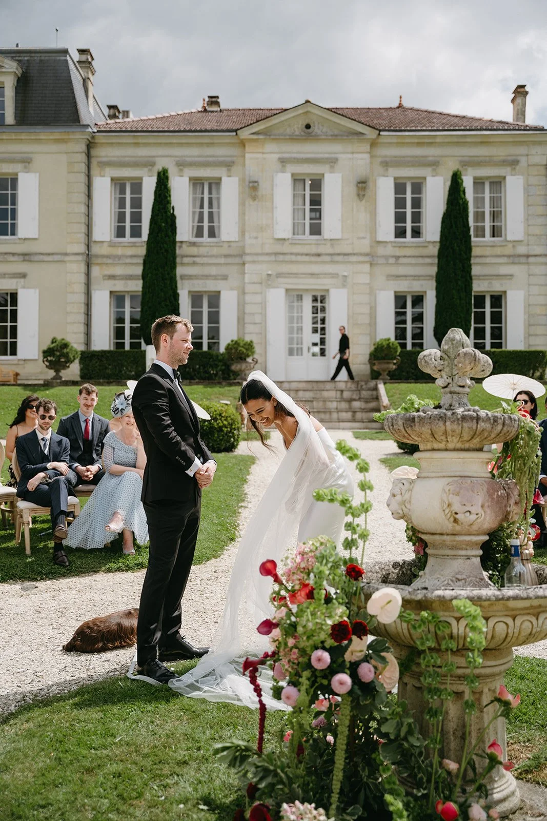 A bride and groom sharing a moment during a wedding ceremony outdoors in front of a large historic mansion, with guests seated nearby. The bride is bowing and laughing, dressed in a white gown and veil, while the groom stands smiling in a black tuxedo. The setting includes a stone fountain and colorful flowers.