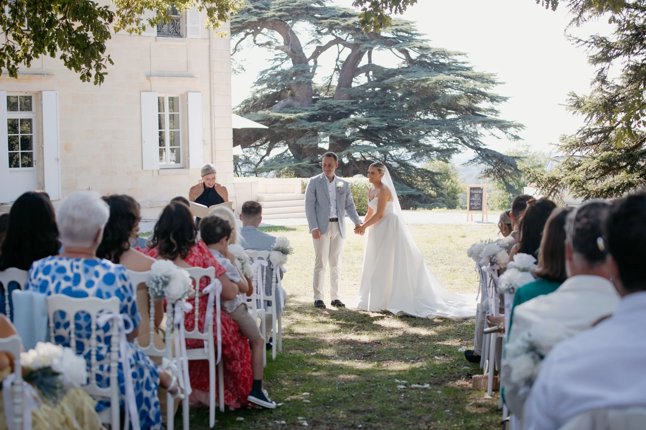 A wedding ceremony outdoors with a bride and groom holding hands, standing in front of officiant, with guests seated on either side, in a garden setting with large trees and a stone building in the background.