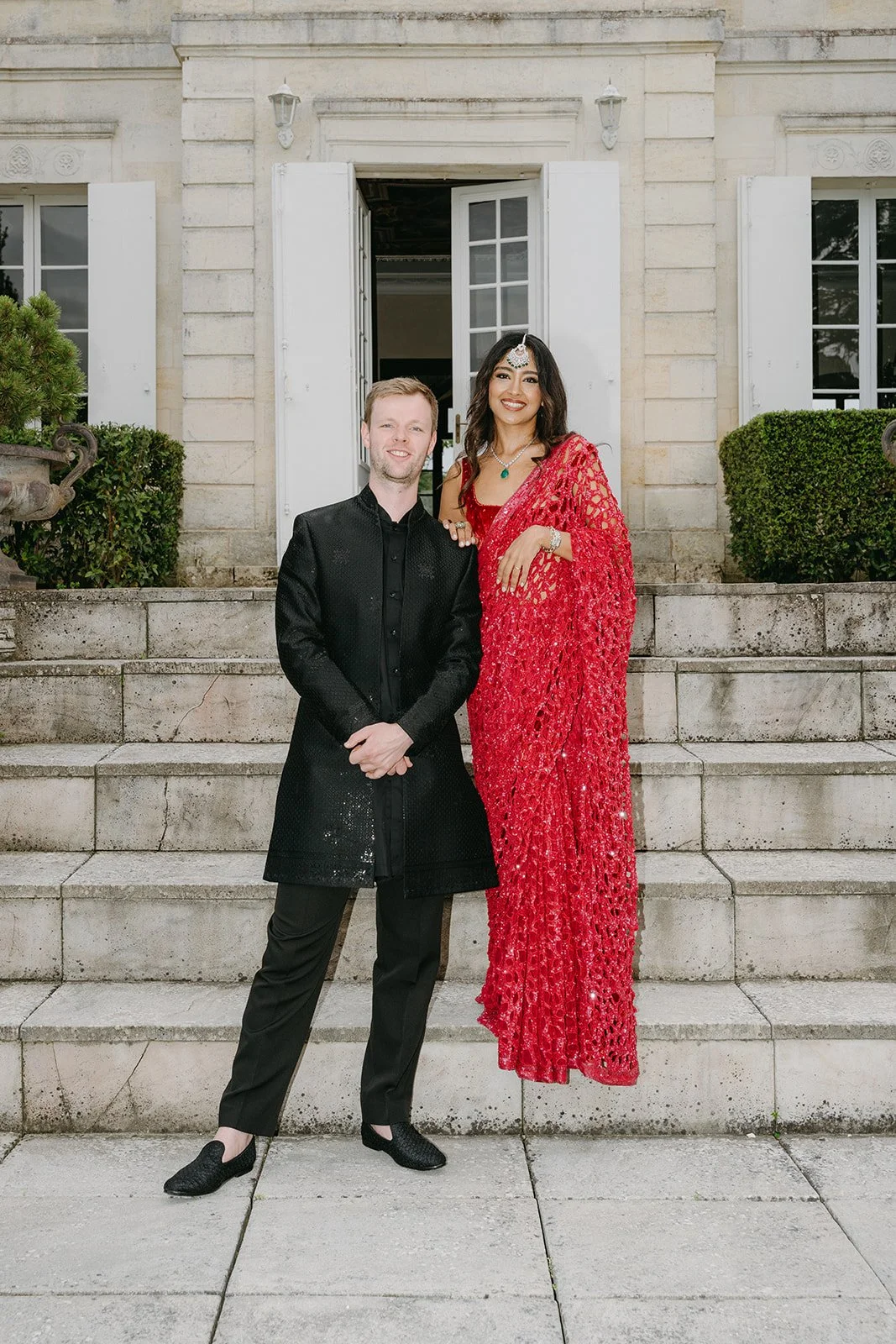A man in a black traditional Indian outfit stands next to a woman in a red saree, both smiling, standing on stone steps outside a building with open white window shutters and greenery.