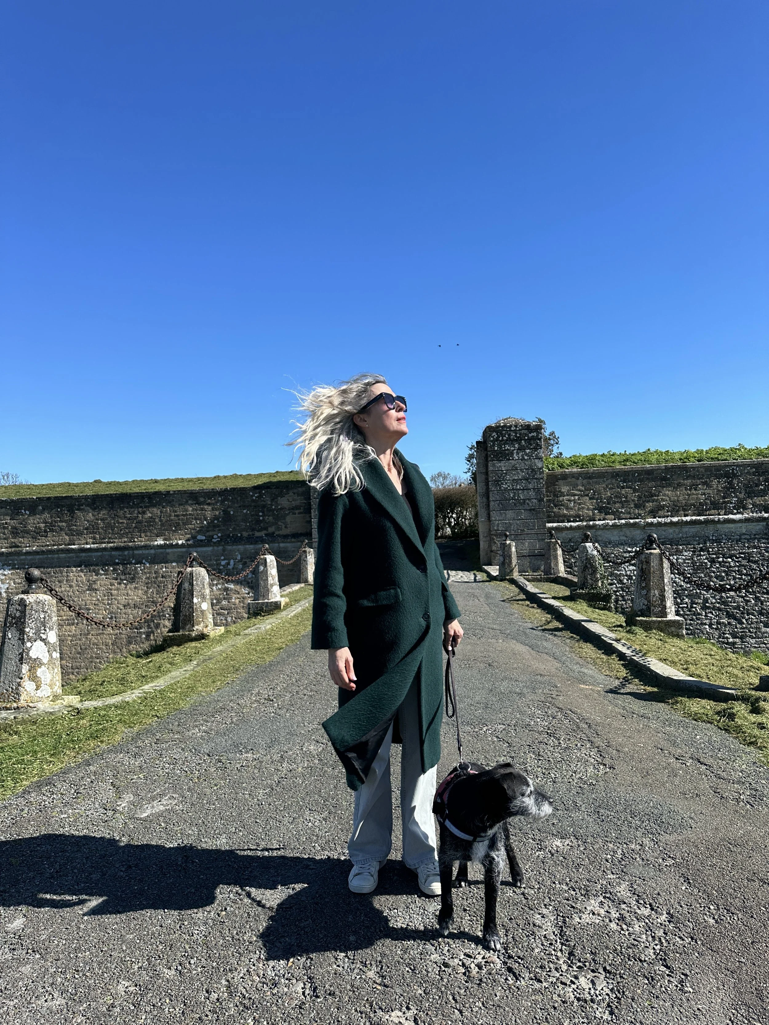 A woman with gray hair wearing sunglasses, a dark green coat, and light-colored pants, standing outdoors on a sunny day with her dog on a leash. The background features stone walls and a bright blue sky.