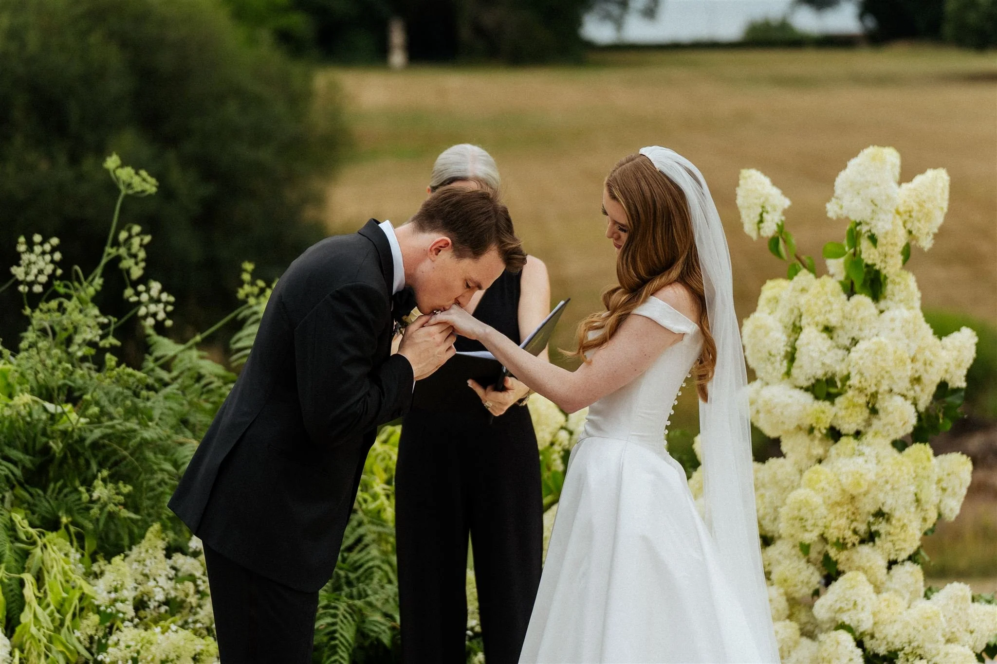 A man in a black tuxedo kisses the hand of a woman in a white wedding dress during a wedding ceremony outdoors, with a woman officiant and large white flowers in the background.