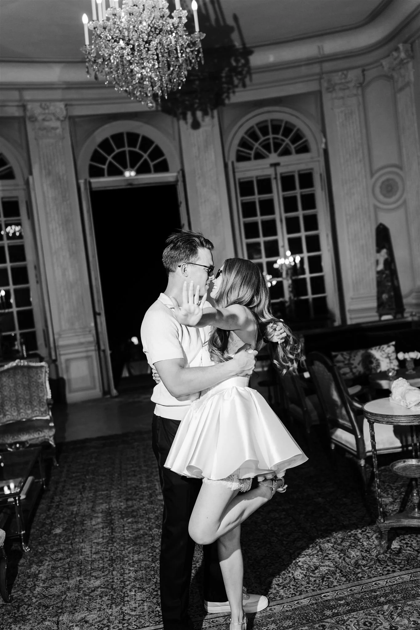 A black and white photo of a couple dancing closely in an elegant, vintage-style room with large arched windows and chandeliers, the woman wearing a short dress and the man wearing glasses and a light-colored shirt.