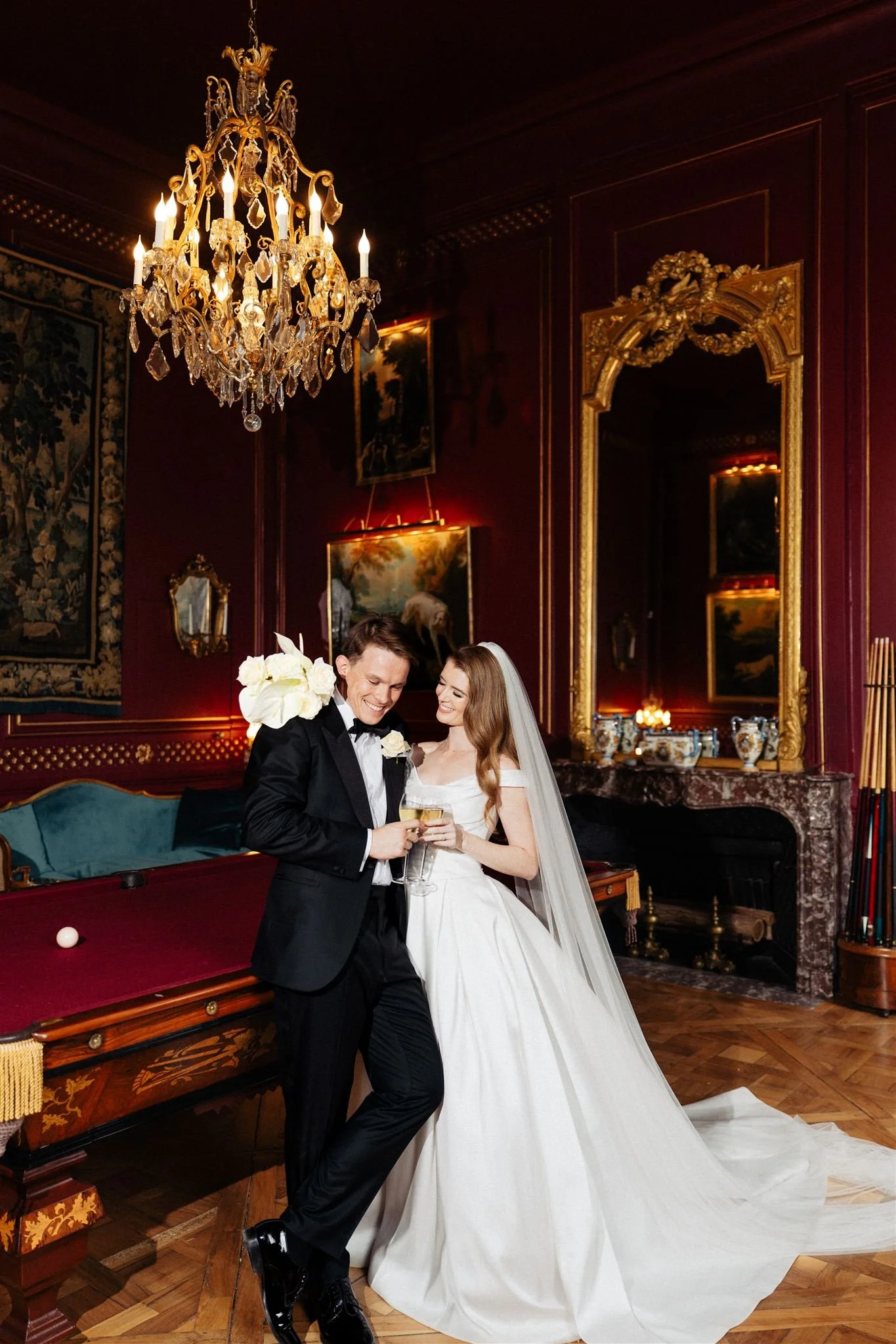 Wedding couple celebrating in an elegant, richly decorated room, with a chandelier, paintings, and ornate gold mirror.