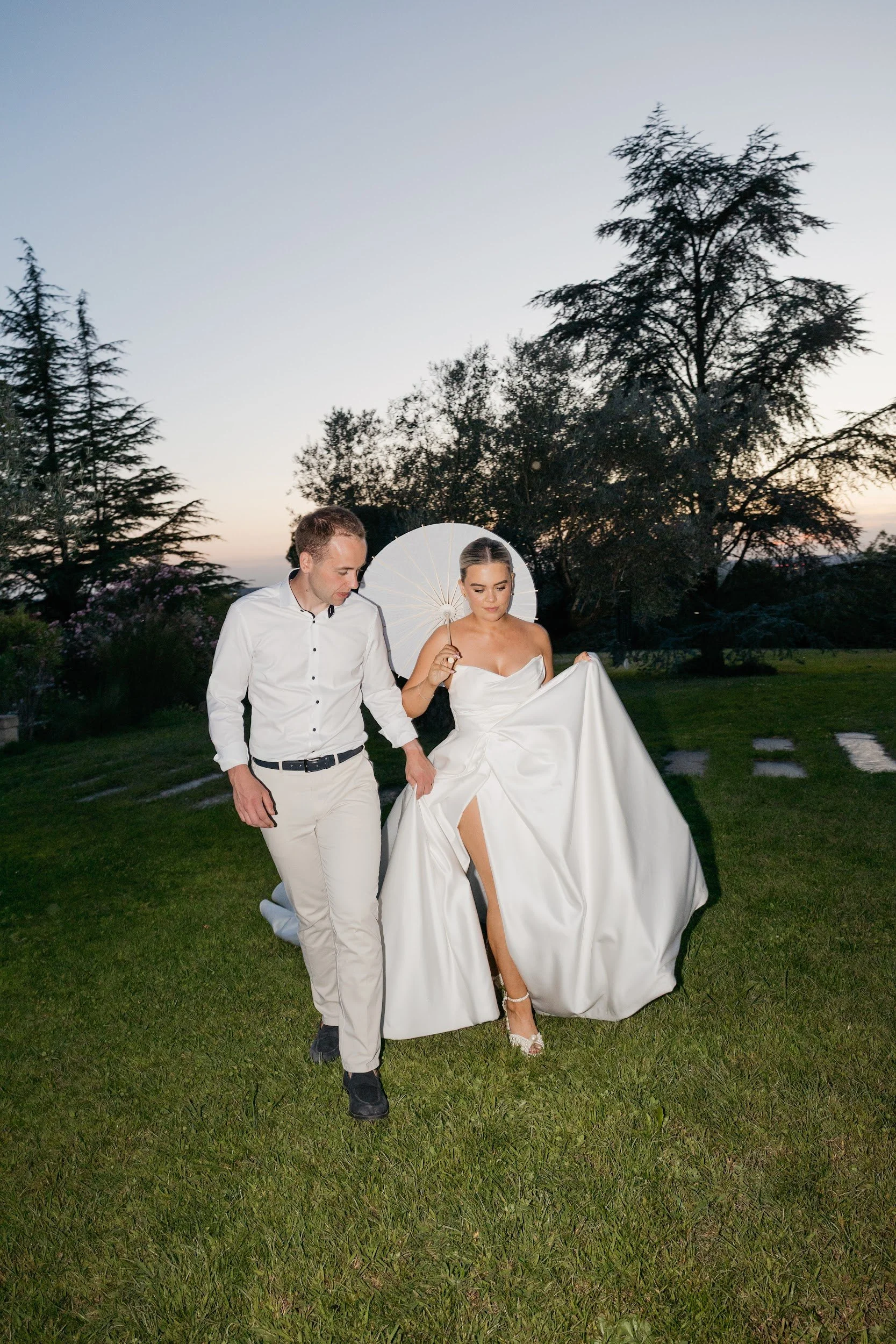 A bride and groom walking on a grassy area during sunset, with the bride holding a white parasol and lifting her wedding dress to reveal her leg.
