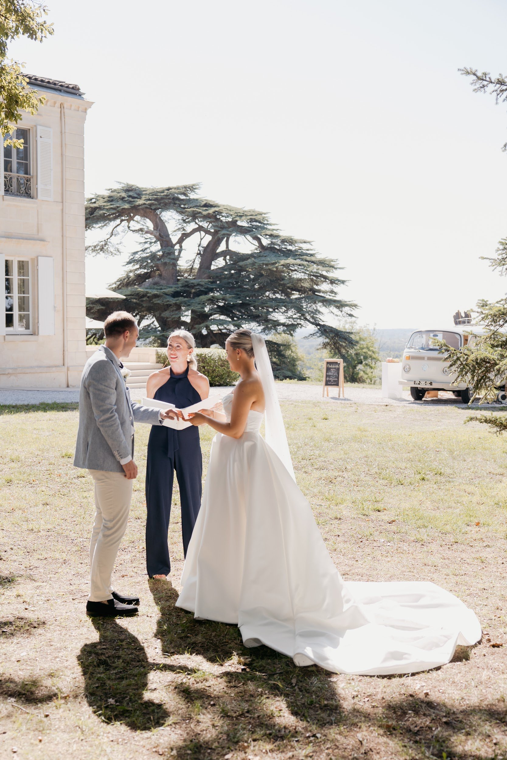 A bride and groom exchanging vows during an outdoor wedding ceremony with an officiant standing between them, on a sunny day with trees and a vintage van in the background.