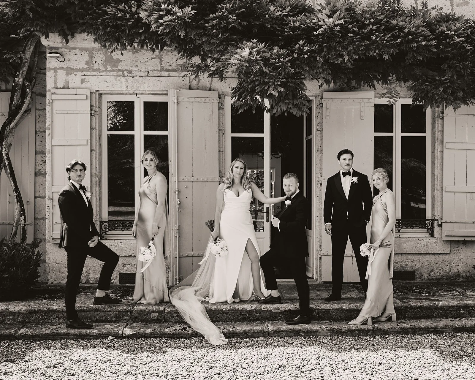 Black and white wedding photo of a bride, groom, and four other women and men standing outside a rustic stone building with open shutters and large windows, under a tree with broad leaves.
