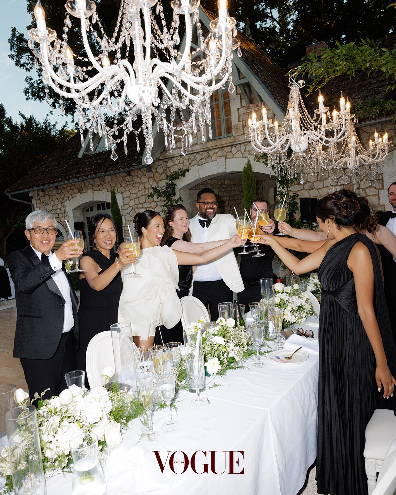 People in formal attire celebrating and raising drinks at an outdoor wedding reception with chandeliers and floral decorations.