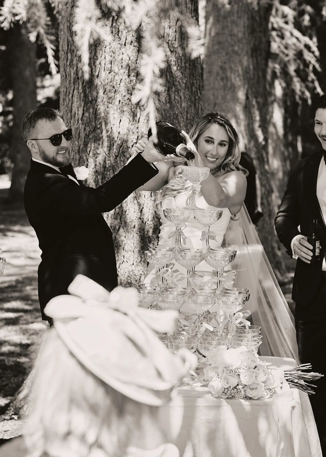 People in wedding attire celebrating outdoors, pouring champagne into a tower of glasses, with a large tree in the background.