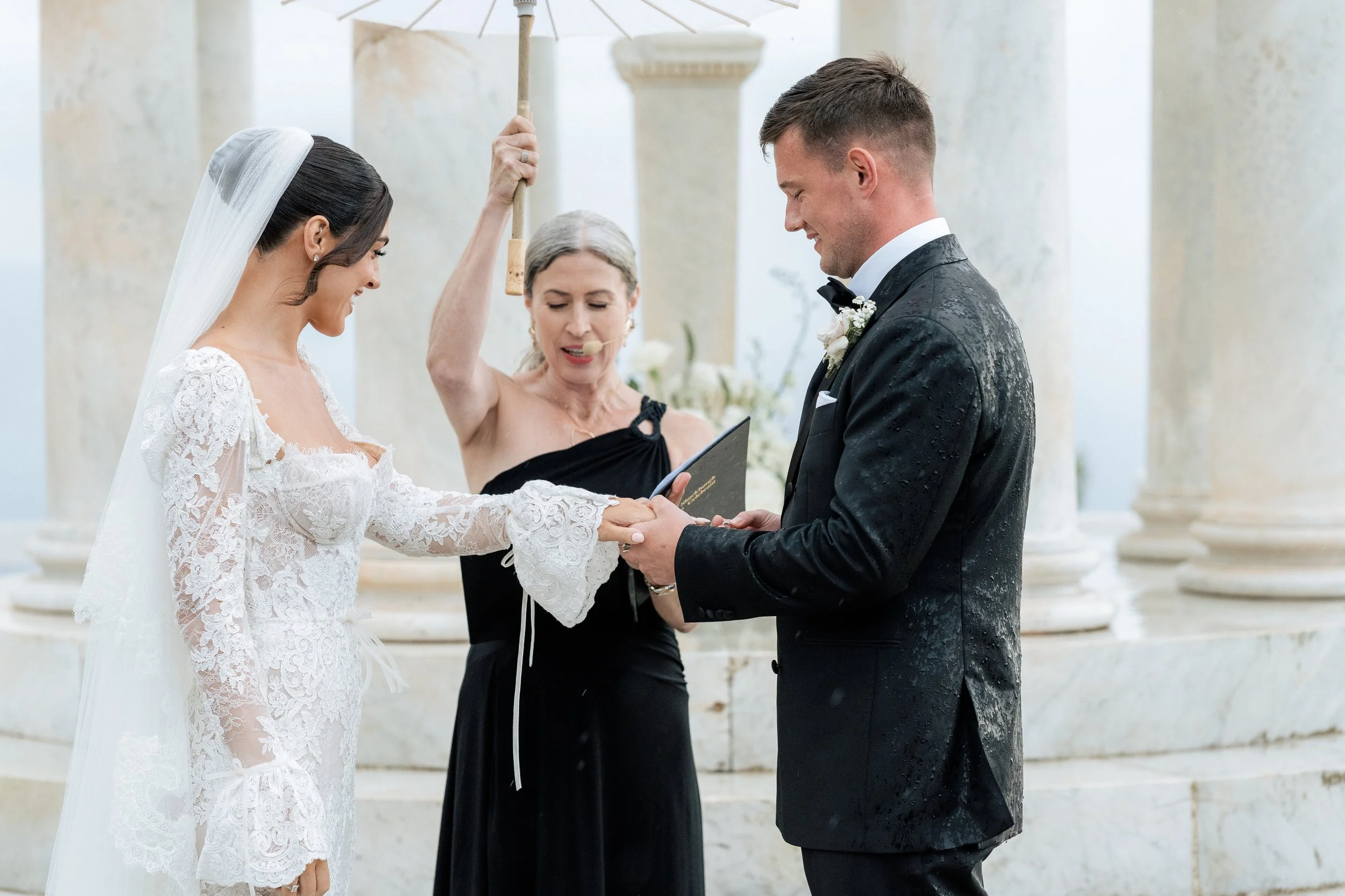 Bride and groom exchanging vows during a wedding ceremony, The Chateau Celebrant holding a book and speaking, set outdoors with columns and flowers.