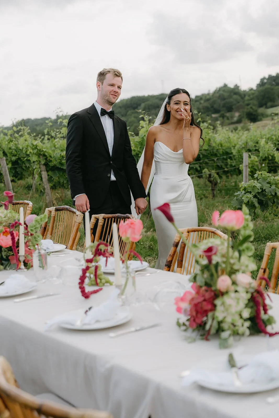 A bride and groom holding hands outdoors during their wedding reception, with the bride smiling and the groom looking at her, surrounded by a decorated table and scenic landscape.