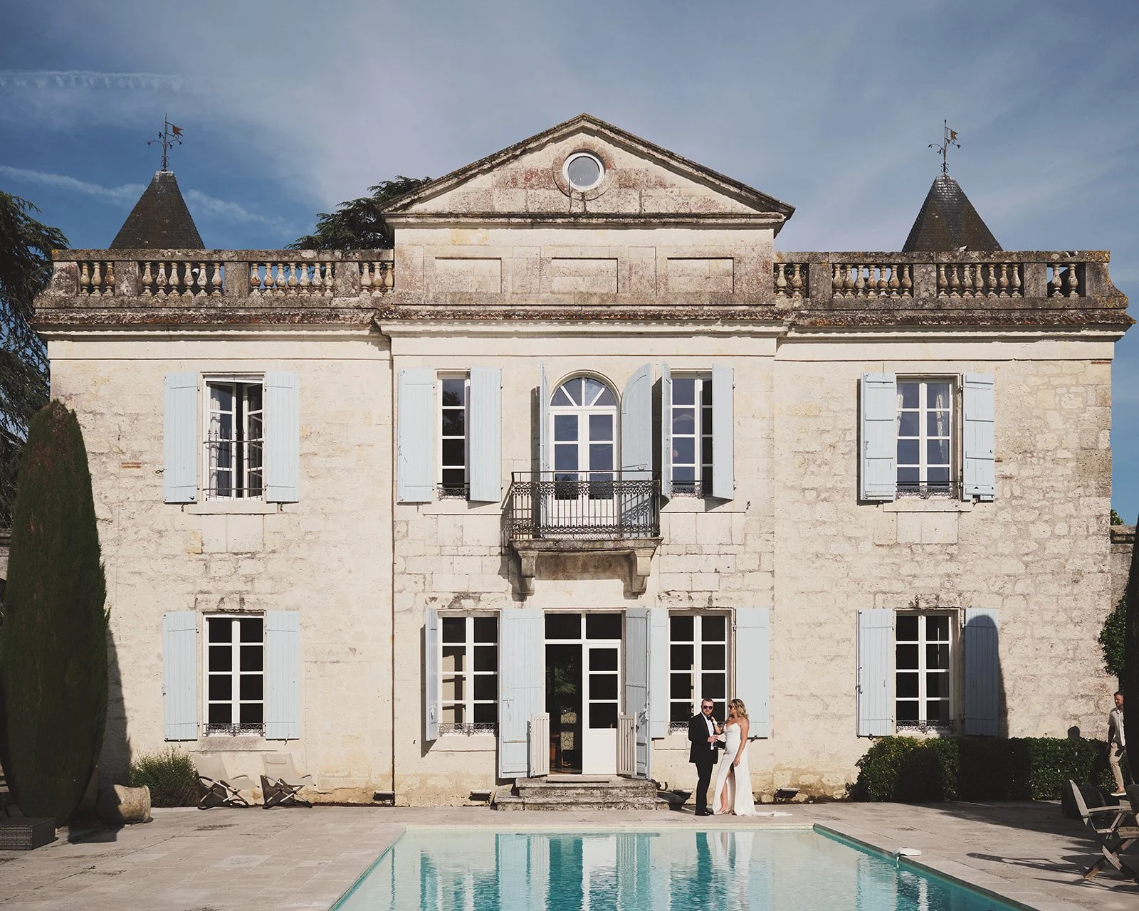 A large, historic mansion with light-colored stone walls and multiple windows with blue shutters, a balcony, and a pool in the foreground. Two people, a man and a woman, are standing near the pool in formal attire, possibly at a wedding.