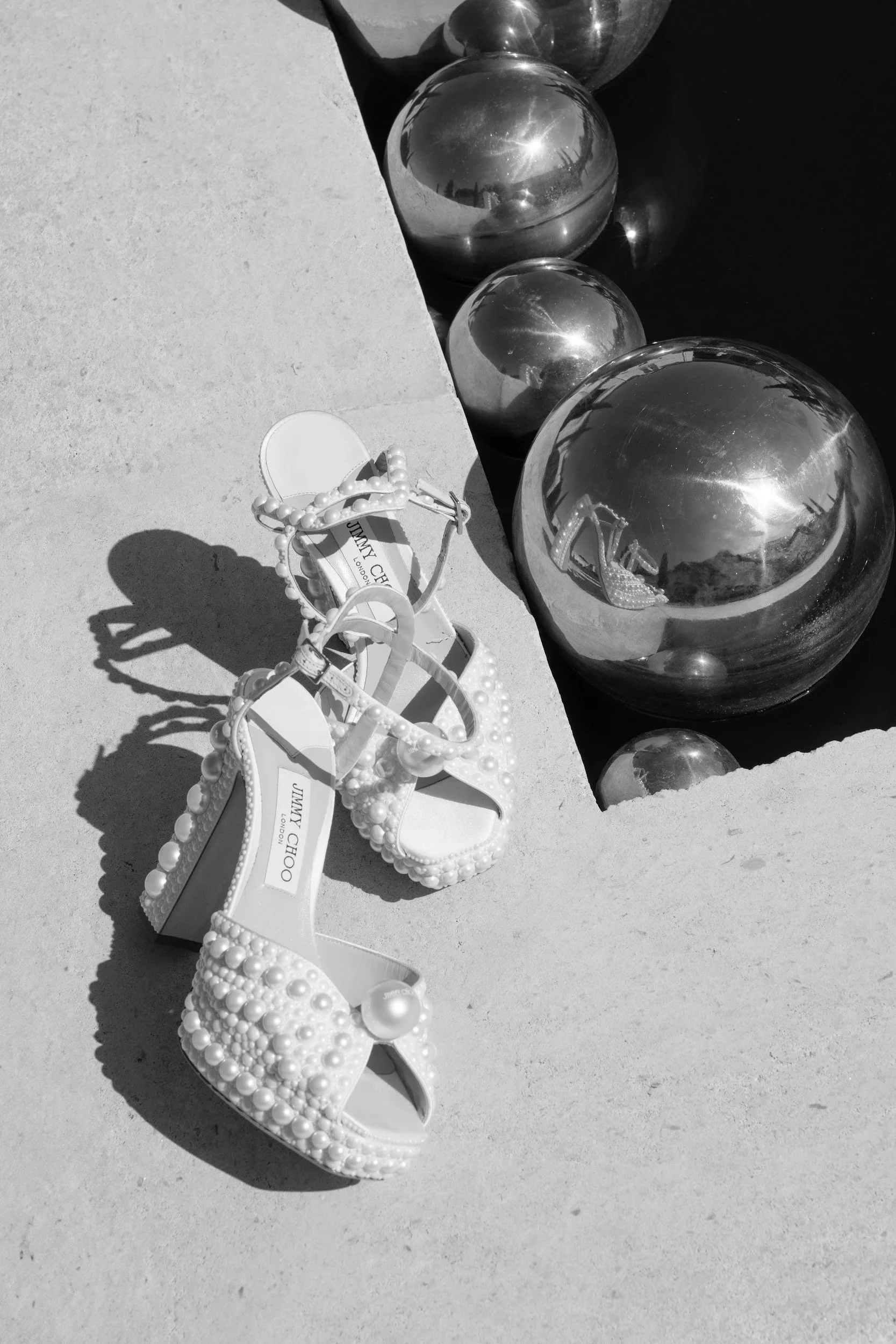 A pair of pearl-embellished high-heeled sandals by Jimmy Choo lies on a concrete surface near reflective metallic spheres of various sizes.