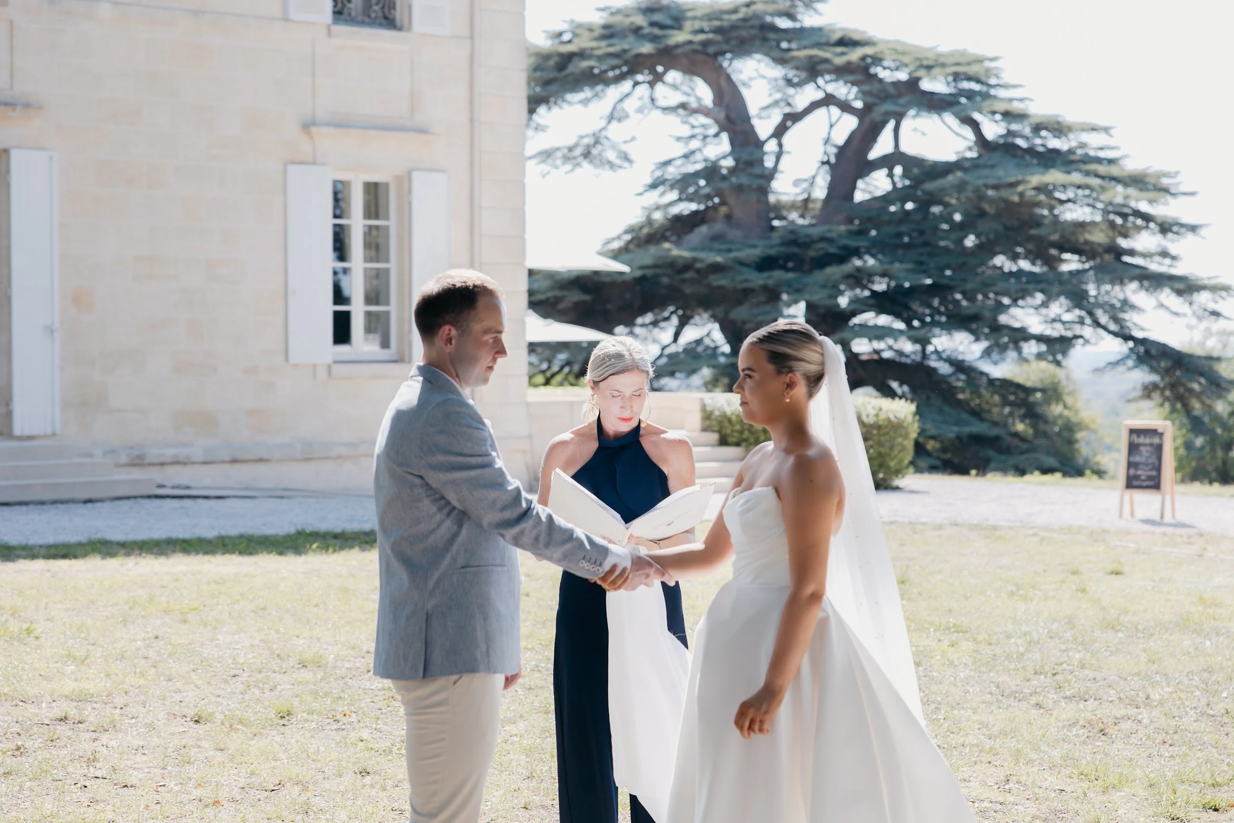 A bride and groom holding hands during their wedding ceremony outdoors, with an celebrant reading from a book against a backdrop of a large tree and a Chateau.