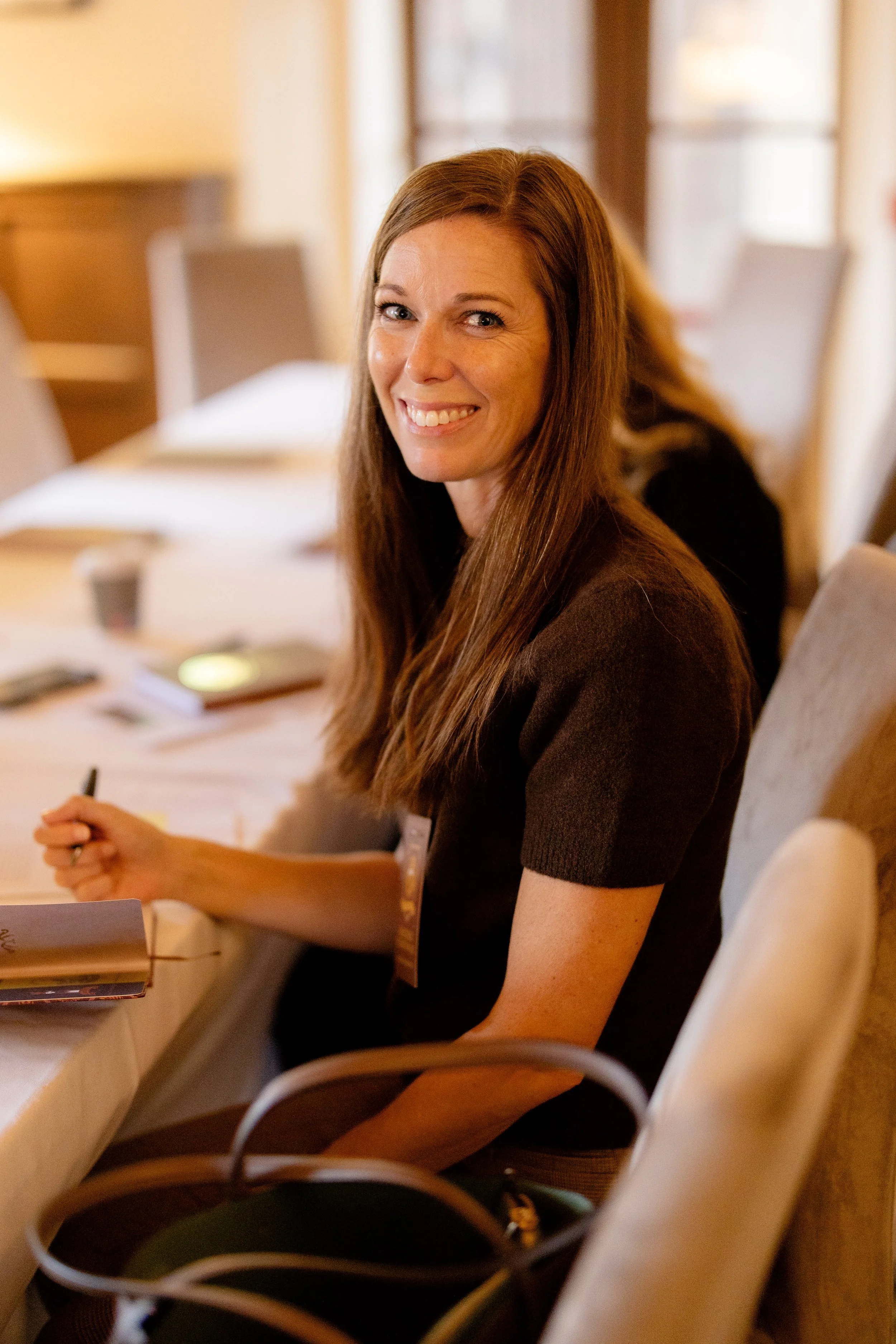 A woman with long brown hair and a dark brown top, smiling and holding a pen, sitting at a conference table in a warmly lit room.