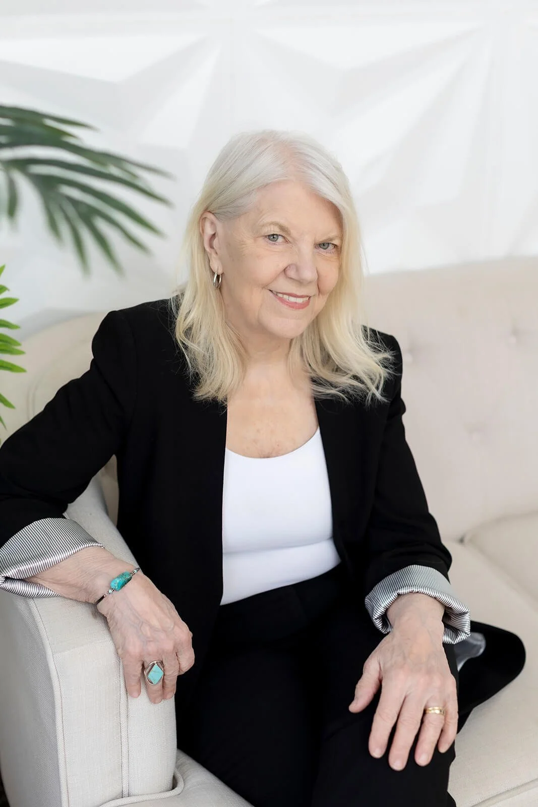 Sharon Kopyc wearing a black blazer over a white top, sitting on a beige sofa with green plants nearby, smiling at the camera.