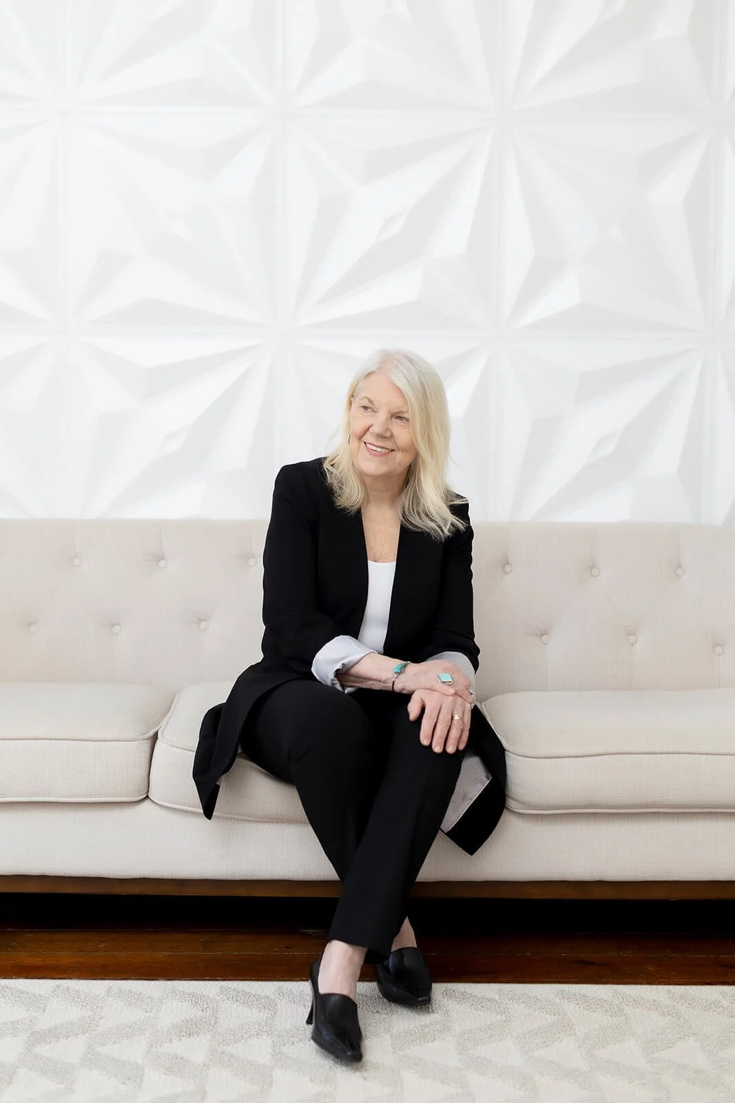 Sharon Kopyc sitting on a beige sofa, smiling and looking to her right, wearing a black suit with a white blouse.