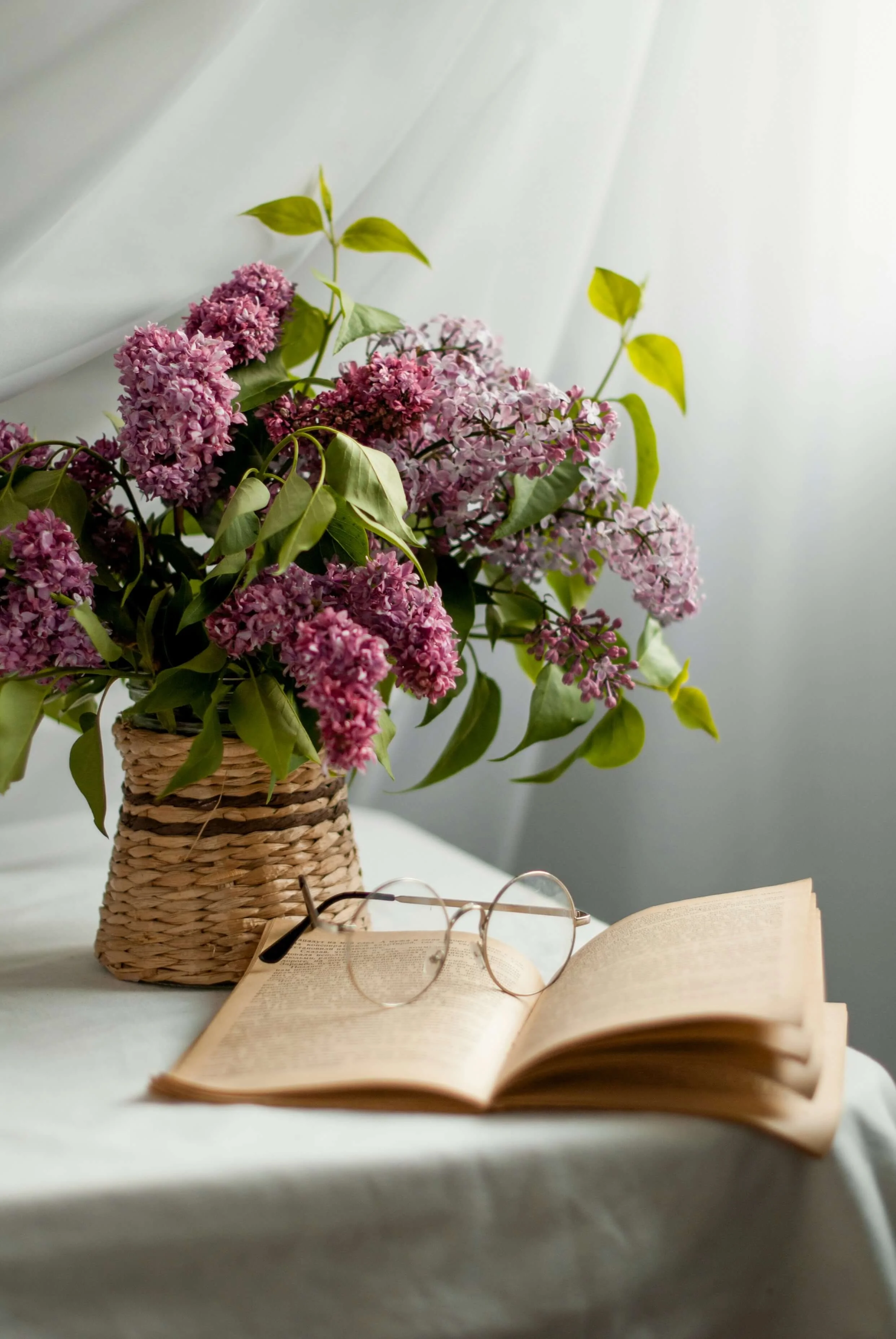 A wicker basket with a bouquet of pink and purple lilac flowers, an open book, and a pair of glasses on a white surface, with a light-colored curtain in the background.