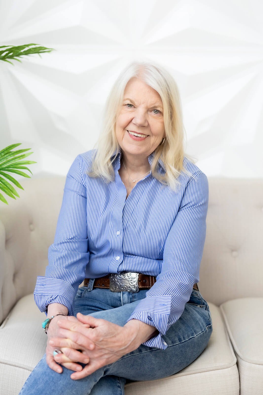 Sharon Kopyc smiling in a friendly post on a beige sofa, wearing a blue and white striped shirt and jeans, with green plants in the background.