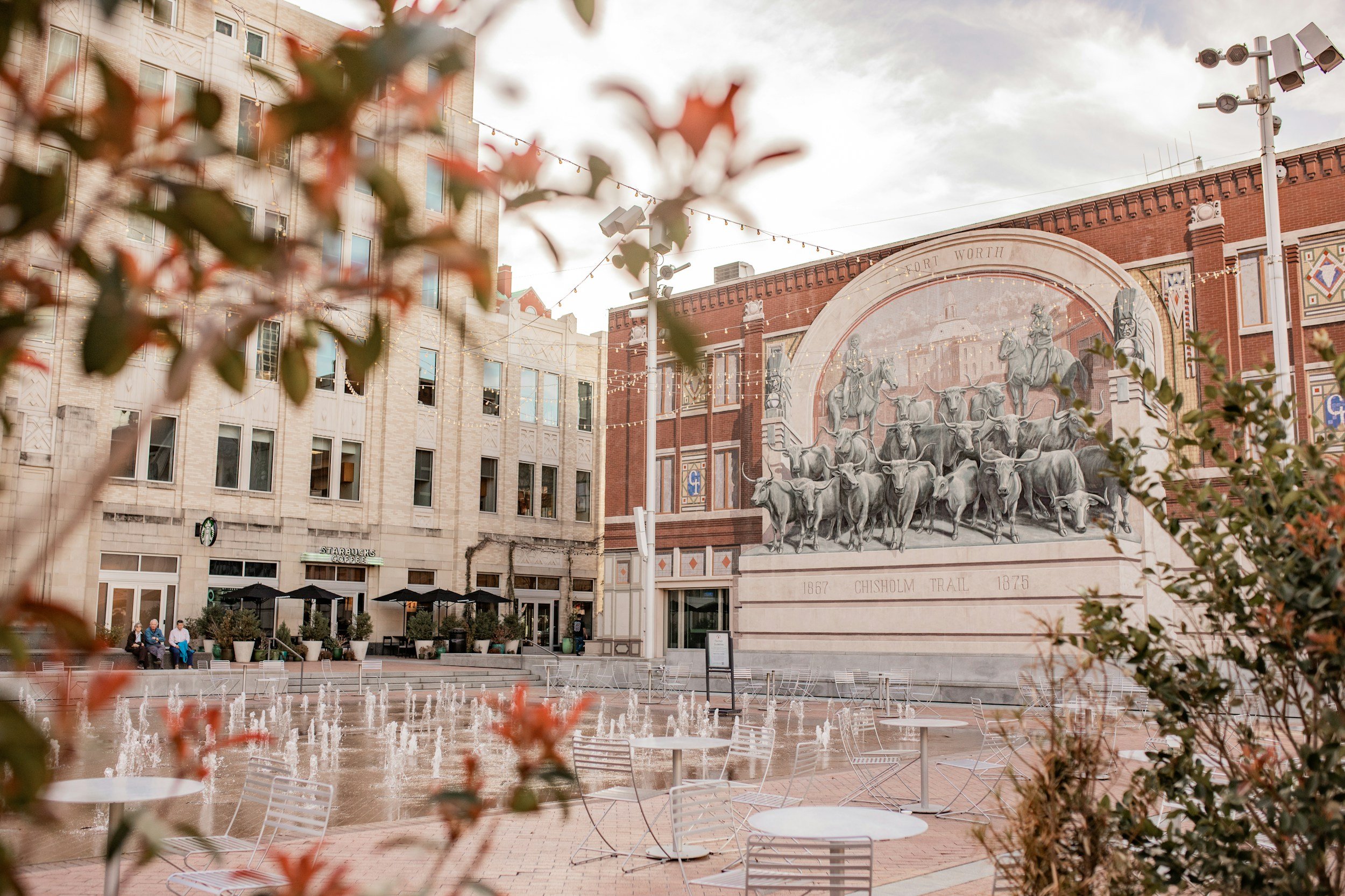 Urban scene with a fountain in a plaza, surrounded by chairs and tables, a mural of cattle on a brick building, and a Starbucks café with outdoor seating. Plants and trees are in the foreground, and there are a few people sitting and walking.