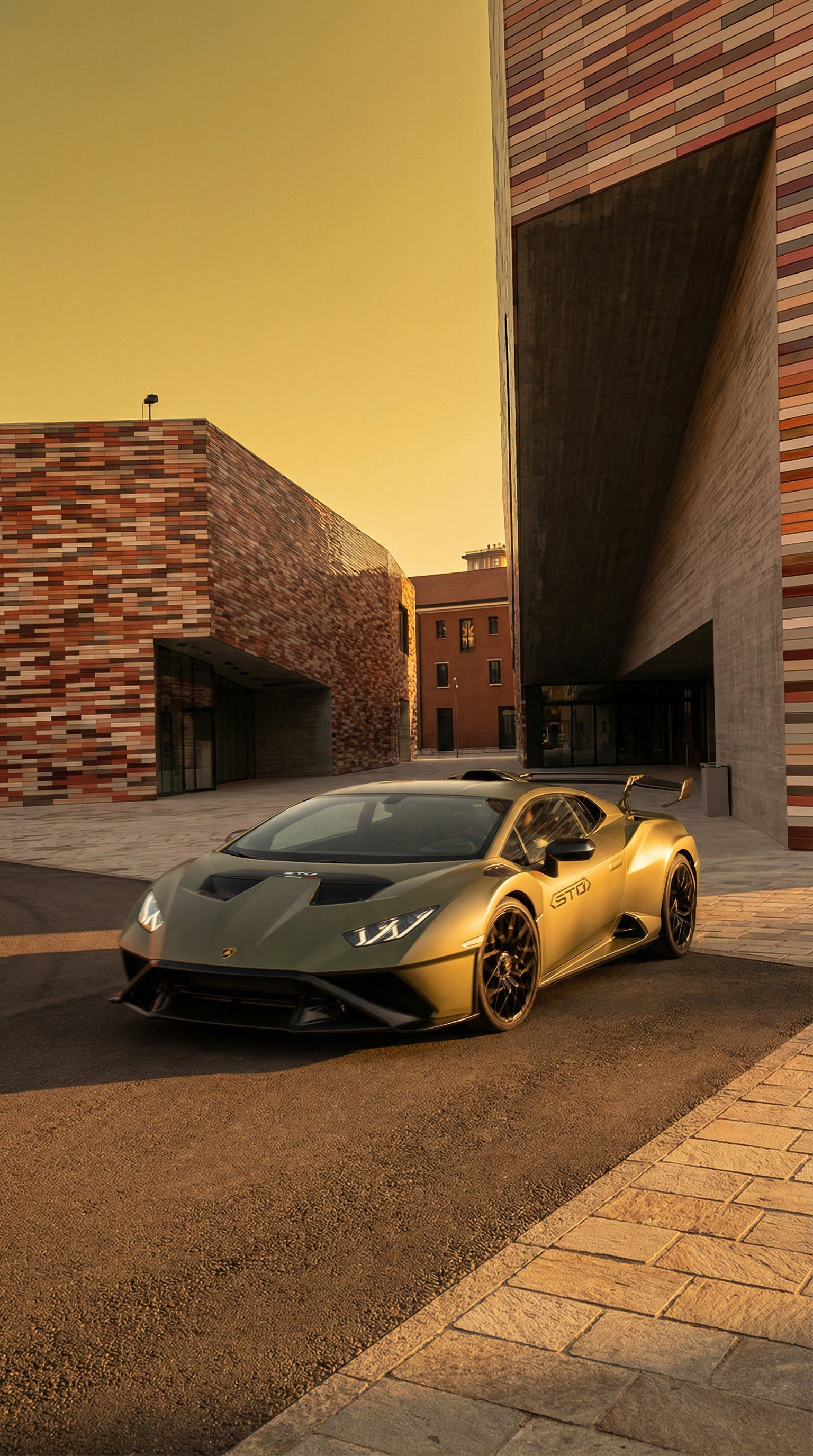 A gold-colored Lamborghini sports car parked in front of modern brick and concrete buildings at sunset.