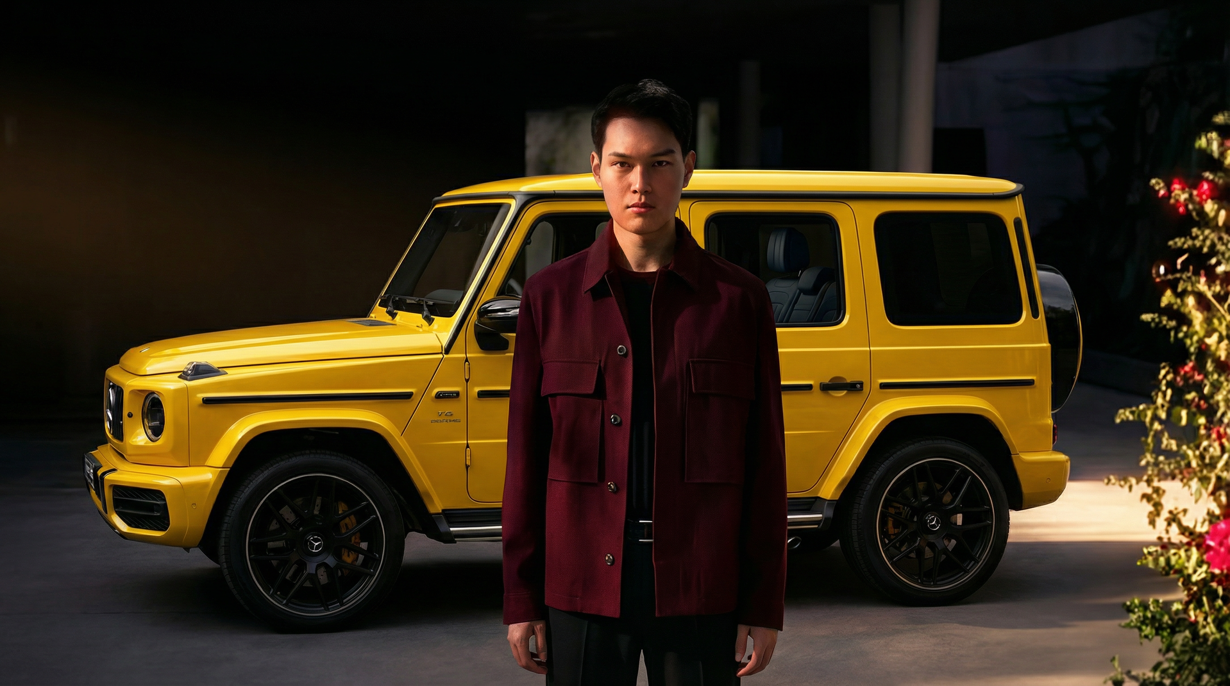 A young man with dark hair wearing a maroon jacket standing in front of a yellow Mercedes-Benz G-Class SUV at night, with flowers visible on the right side.