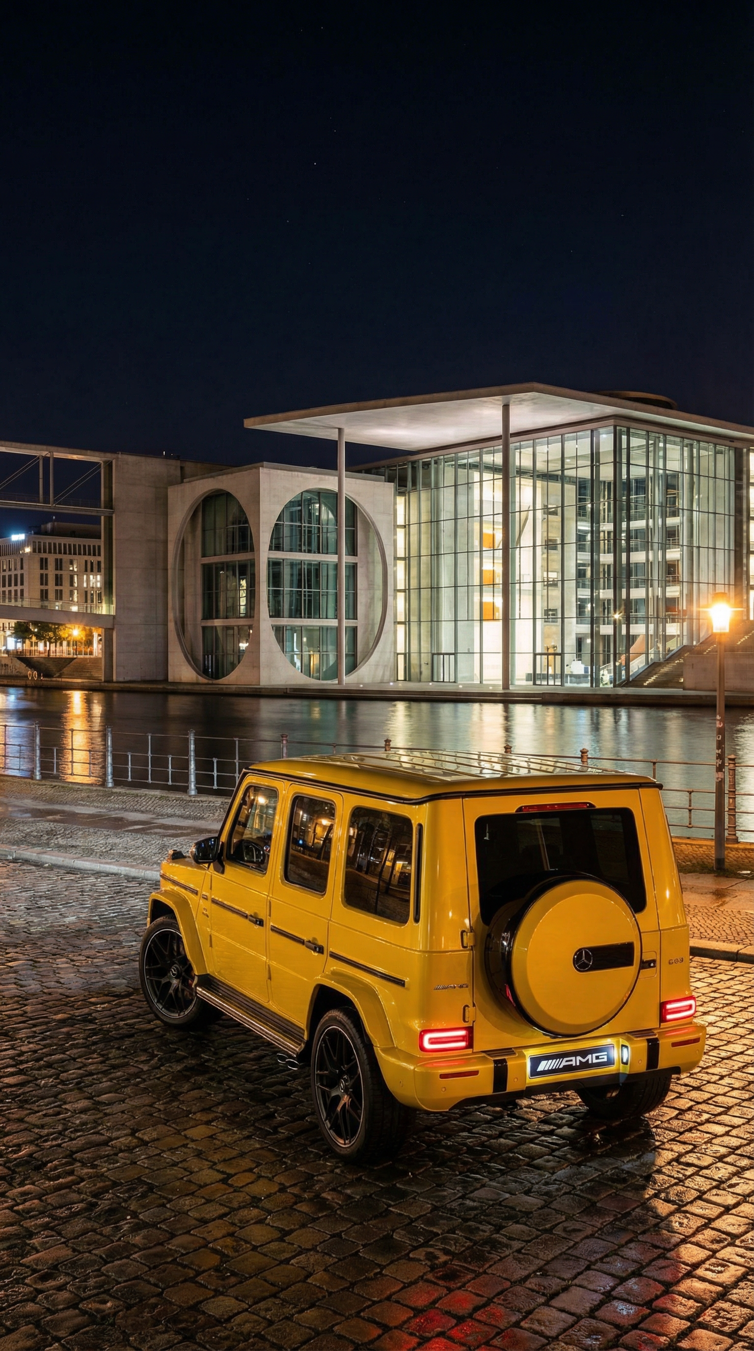 Nighttime photo of a bright yellow Mercedes-Benz G-Class SUV parked on a cobblestone street near a modern building with large glass windows and artful architectural details, reflecting on the water.