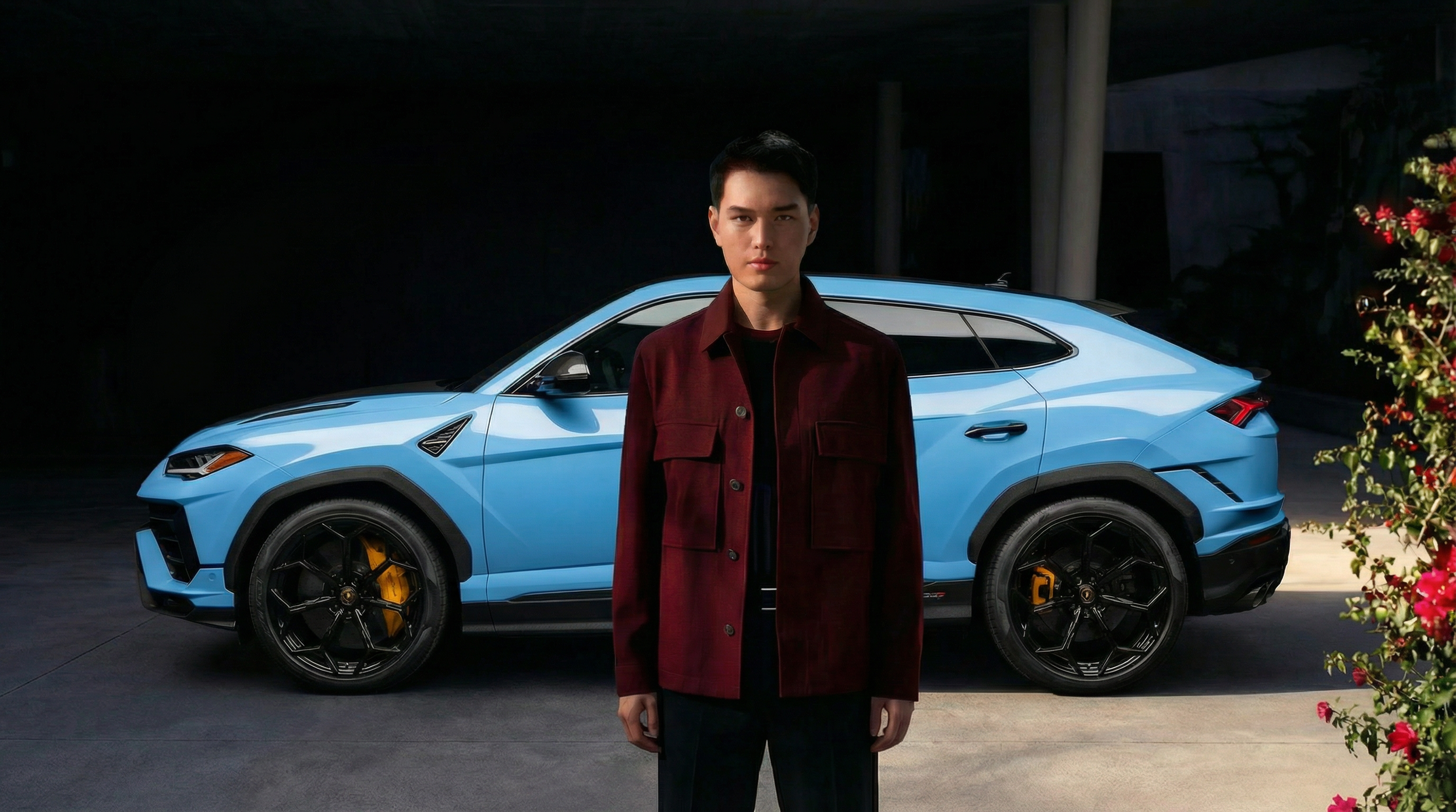 A young man with black hair and a serious expression stands in front of a blue sports SUV, wearing a dark red jacket and black pants. The setting appears to be outdoors at night, with some pink and red flowers visible on the right side of the image.