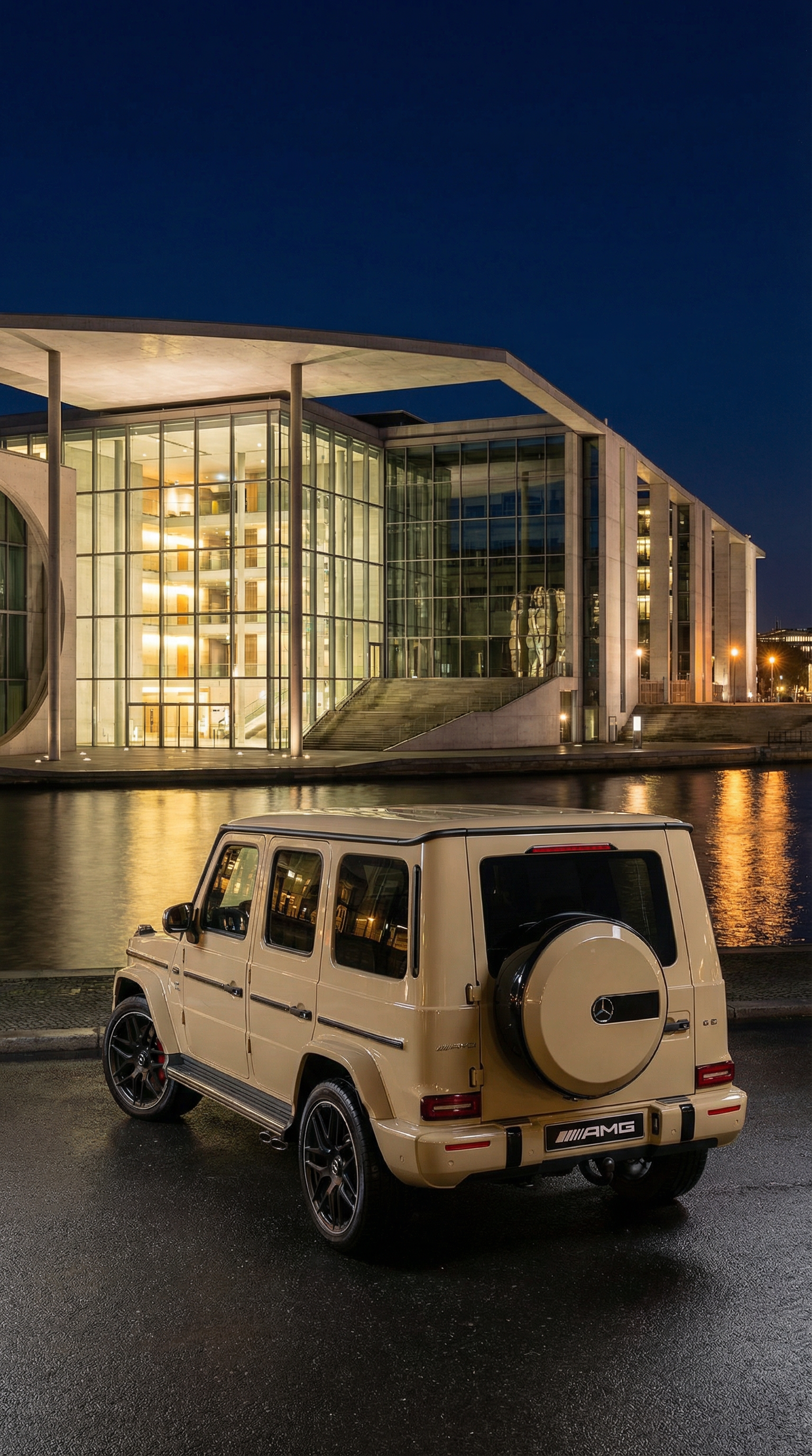 A beige Mercedes-Benz G-Class SUV parked in front of a modern glass building at night, with water reflecting the building's lights.