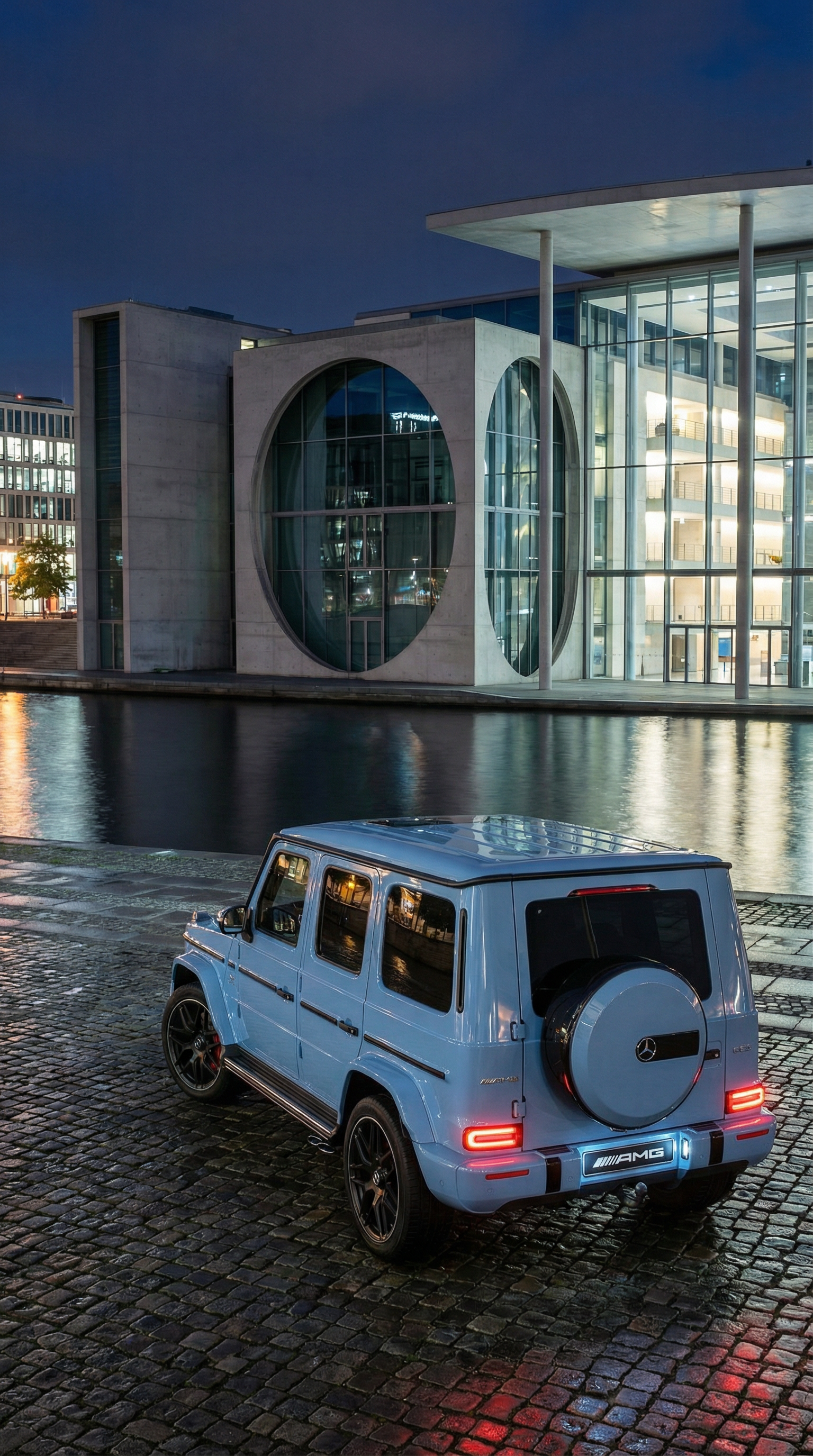 A blue Mercedes-Benz G-Class SUV parked on a cobblestone street at night, with modern glass and concrete buildings reflecting in the water nearby.
