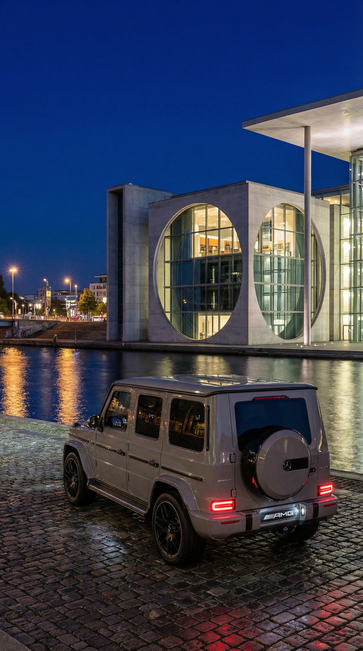A Mercedes-Benz G-Class SUV parked on cobblestone near a waterway in front of a modern building with large circular window design at night.