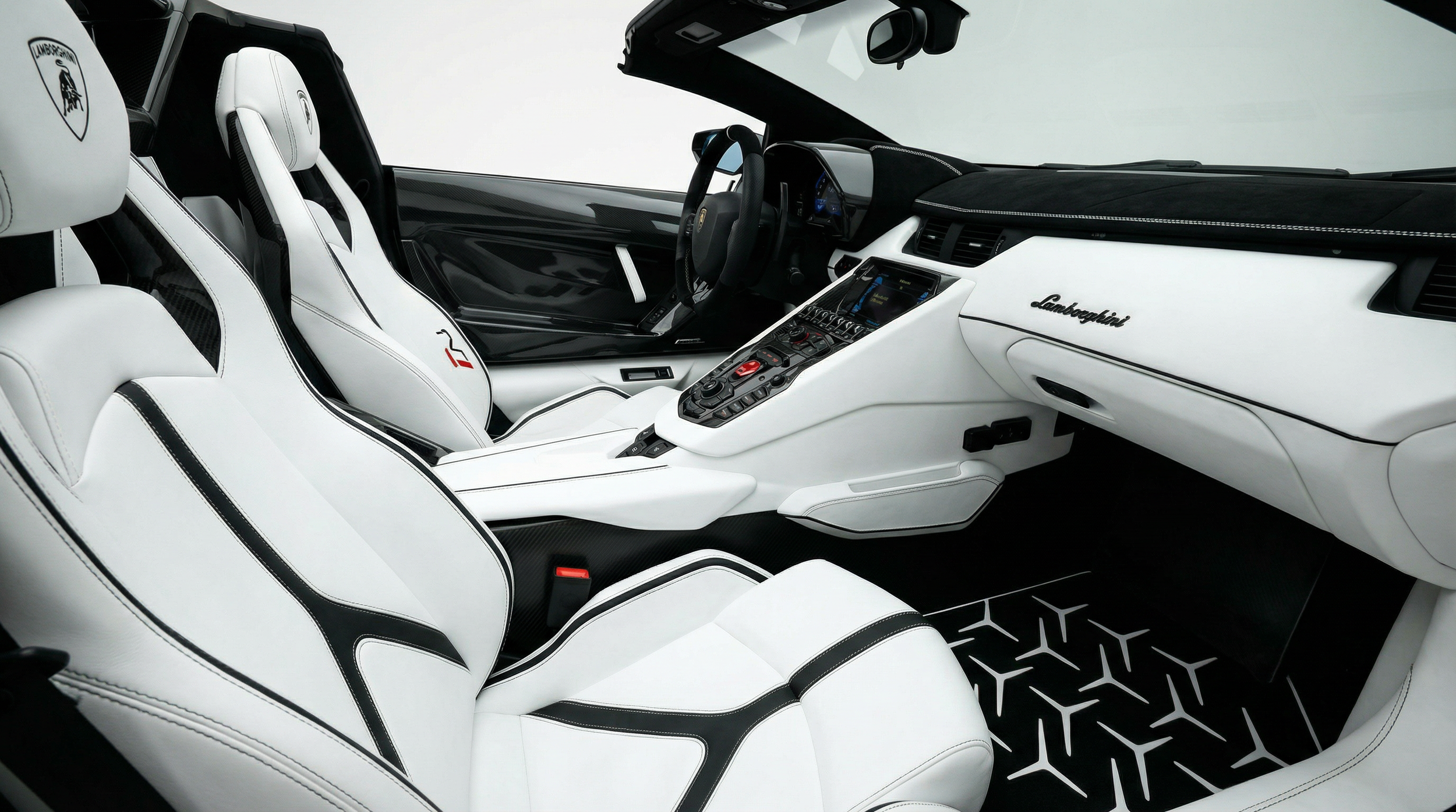 Interior of a white Lamborghini sports car with white leather seats featuring black accents. The dashboard is white with the Lamborghini logo, and the steering wheel is black. The center console has numerous buttons and controls, with a digital display screen visible. The flooring has a black and white geometric pattern.