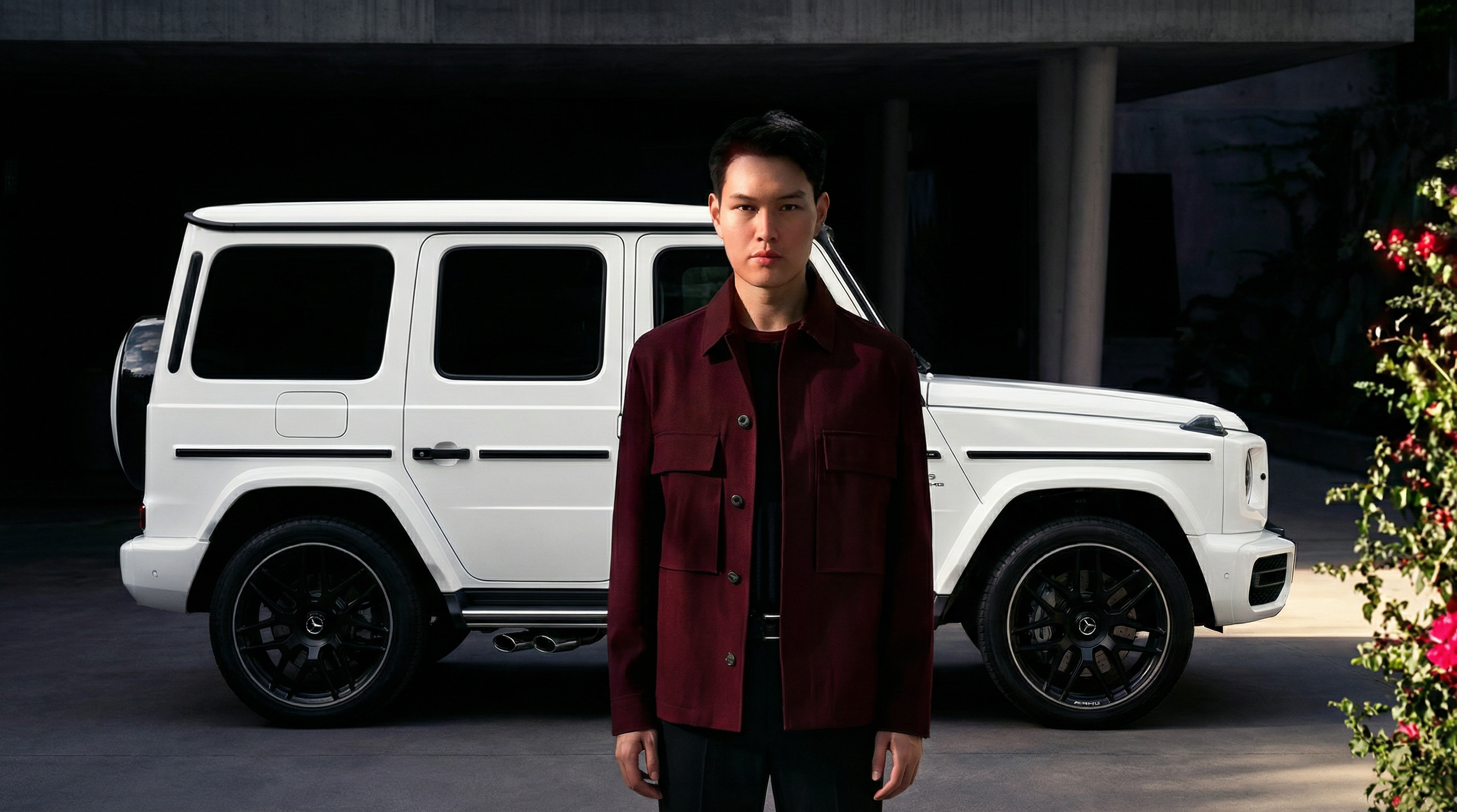 A young man with short black hair standing in front of a white Mercedes-Benz G-Class SUV, wearing a dark red jacket over a black shirt, outdoors during daytime.