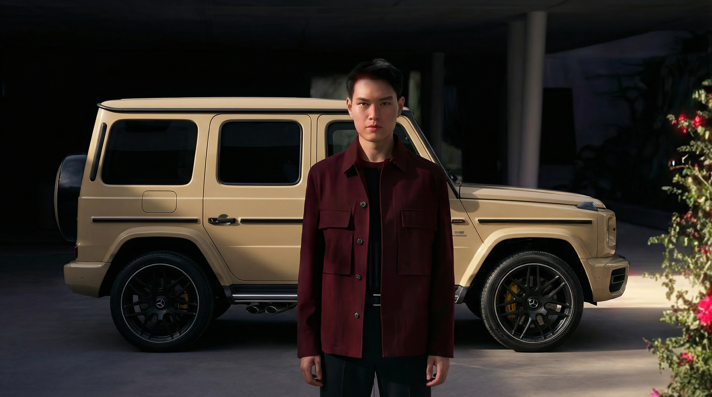 A young man standing in front of a beige Mercedes-Benz G-Class SUV under a bridge or parking structure, with a flower bush on the right side.