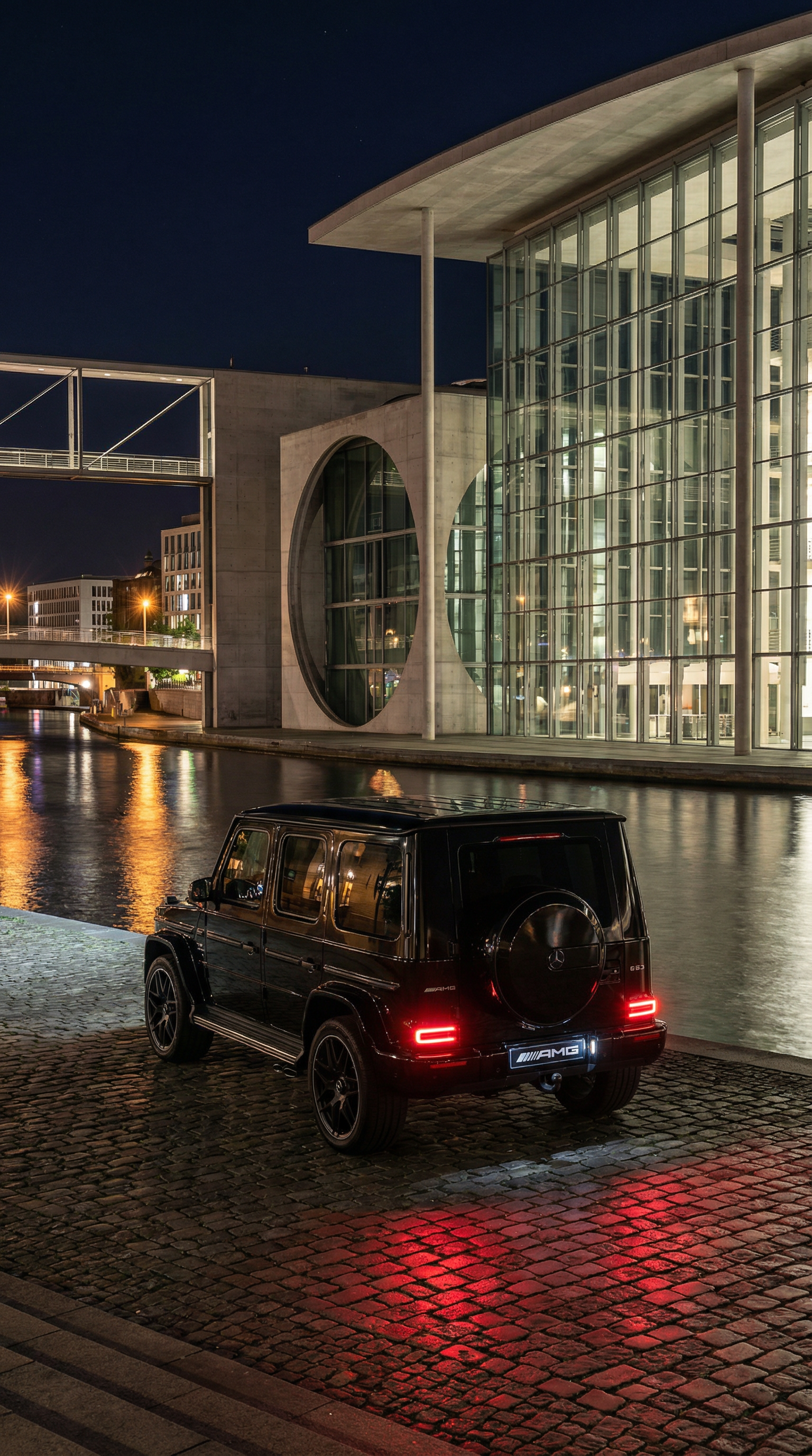Nighttime cityscape with modern glass building reflecting lights, and a black Mercedes-Benz G-Class SUV parked on cobblestone near a river.