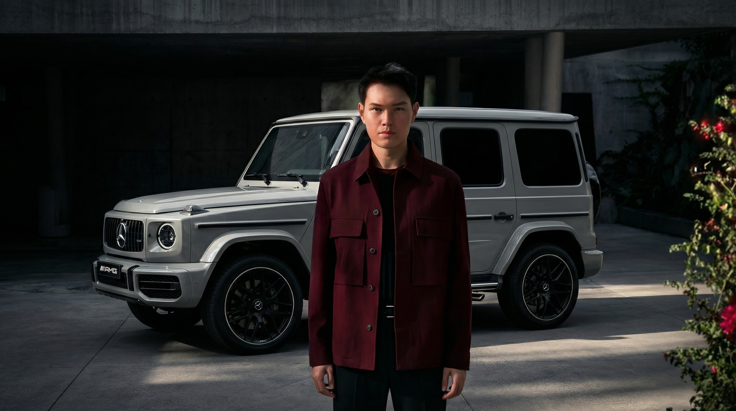 A young man with dark hair wearing a dark red jacket standing in front of a silver Mercedes-Benz G-Class SUV in an outdoor urban setting.