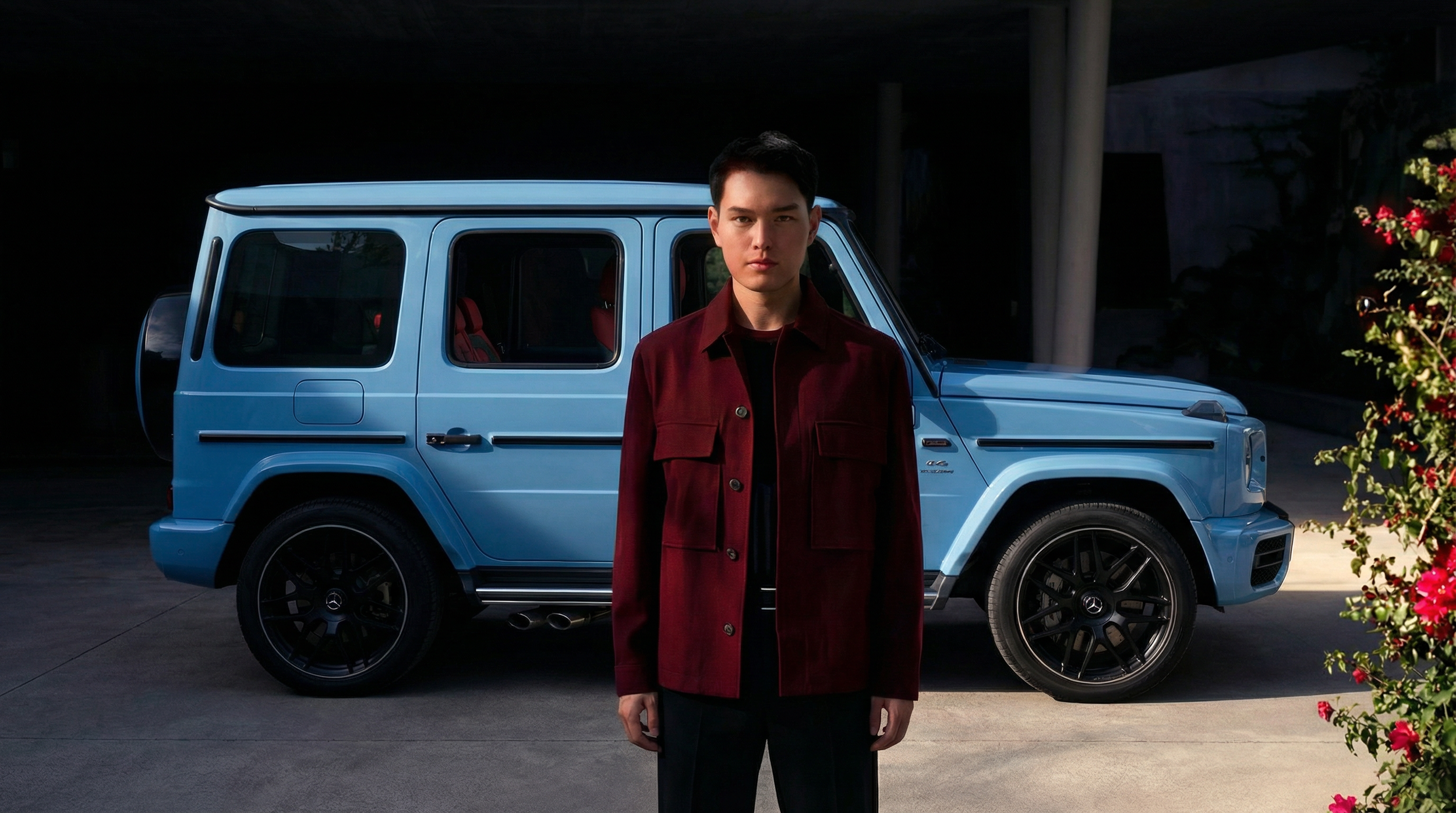 A young man stands in front of a blue Mercedes-Benz G-Class SUV, outdoors at night, with a dark background and some pink flowers visible on the right side.