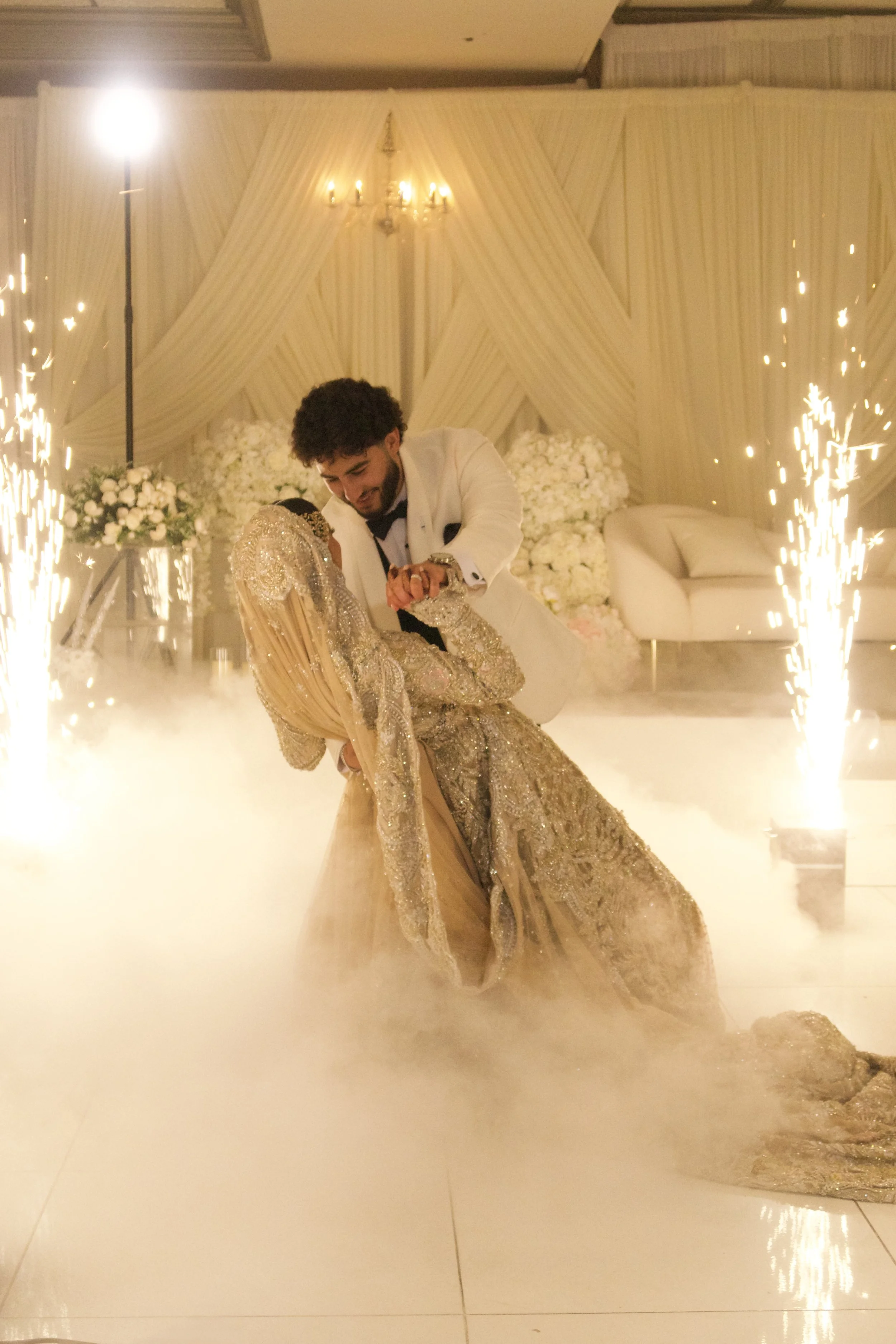 A bride and groom dancing at a wedding with fireworks and fog effects, decorated with white flowers and drapery, in a luxurious venue.