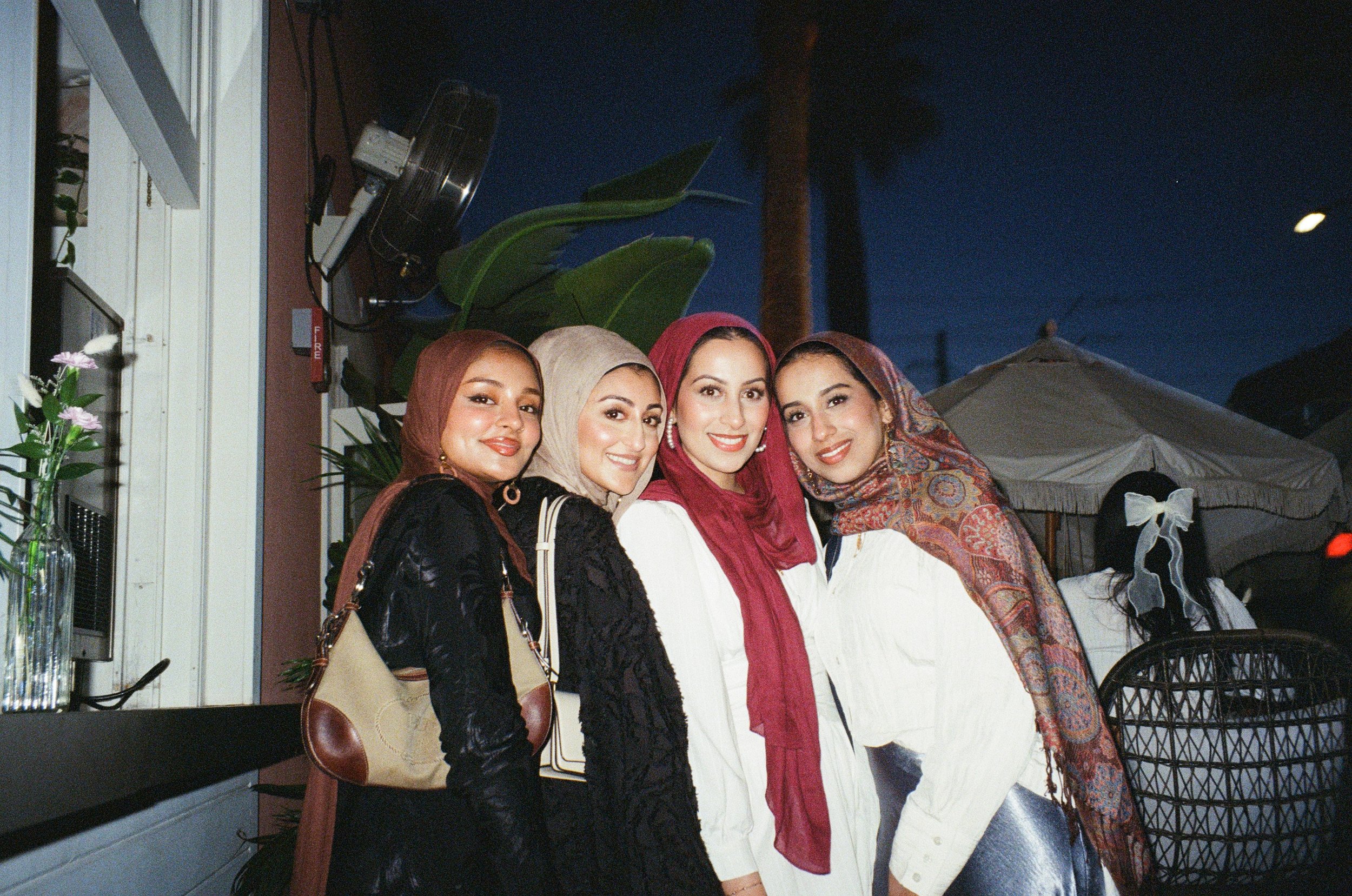 Four women wearing headscarves smiling and posing together at an outdoor evening gathering.