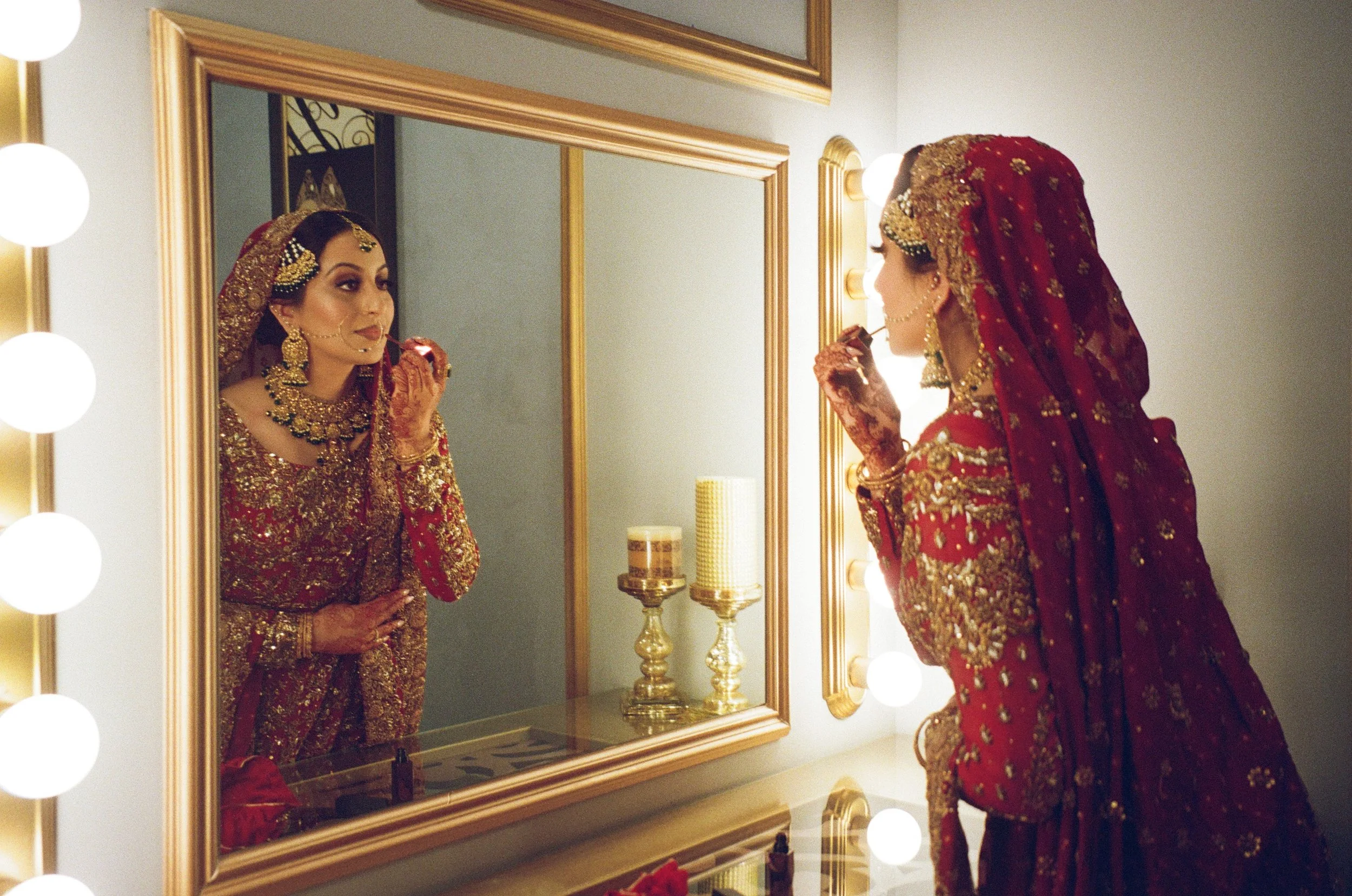A bride dressed in red and gold traditional wedding attire, standing in front of a mirror while applying lipstick, with her reflection visible.